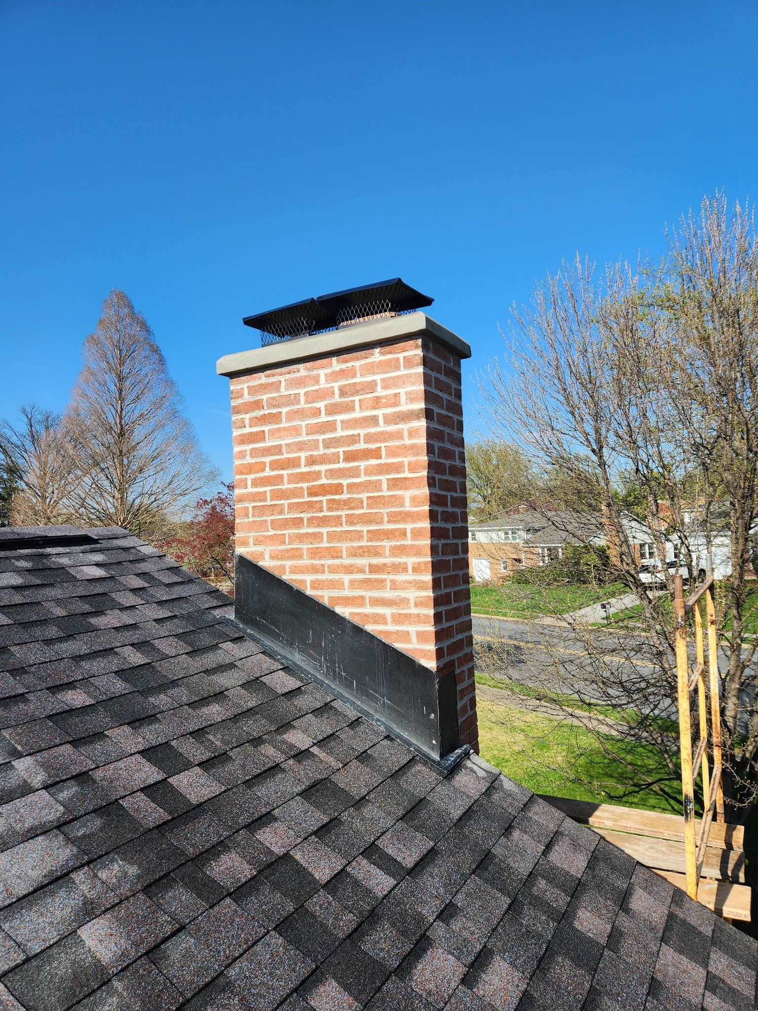 Brick chimney on a dark shingle roof against a blue sky with trees in the background.