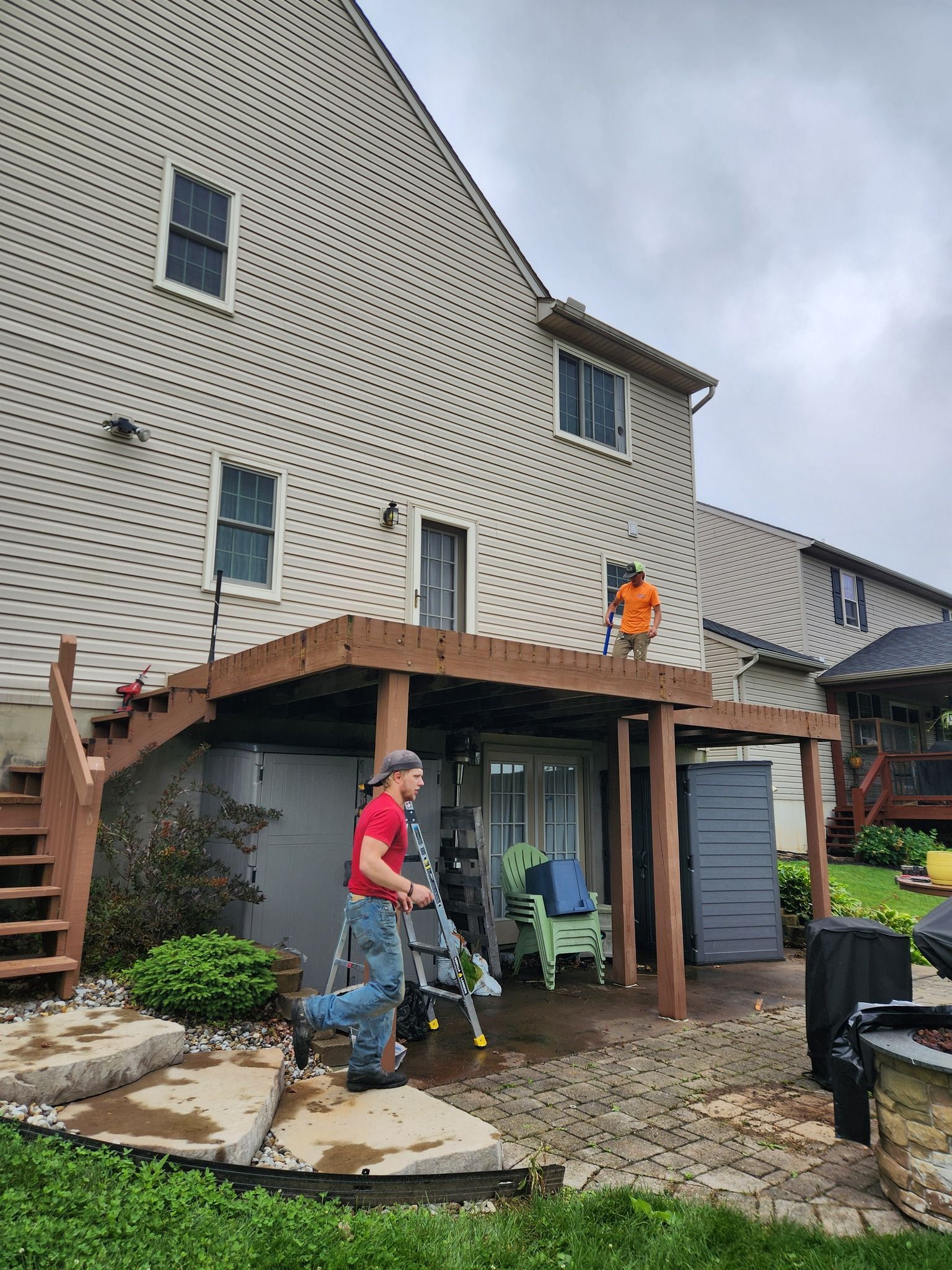 Two men working on a wooden deck attached to a house with a cloudy sky. One on the deck, one on the ground.