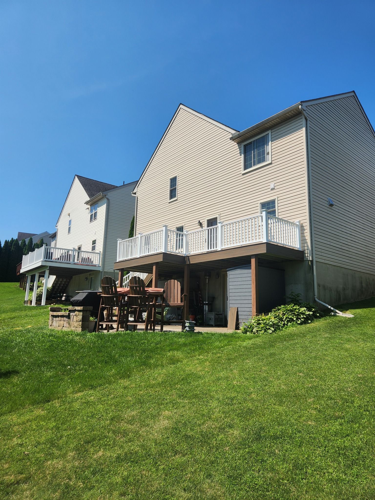 Backyards of two-story houses. One has a deck, the other a patio. Green grass, blue sky.