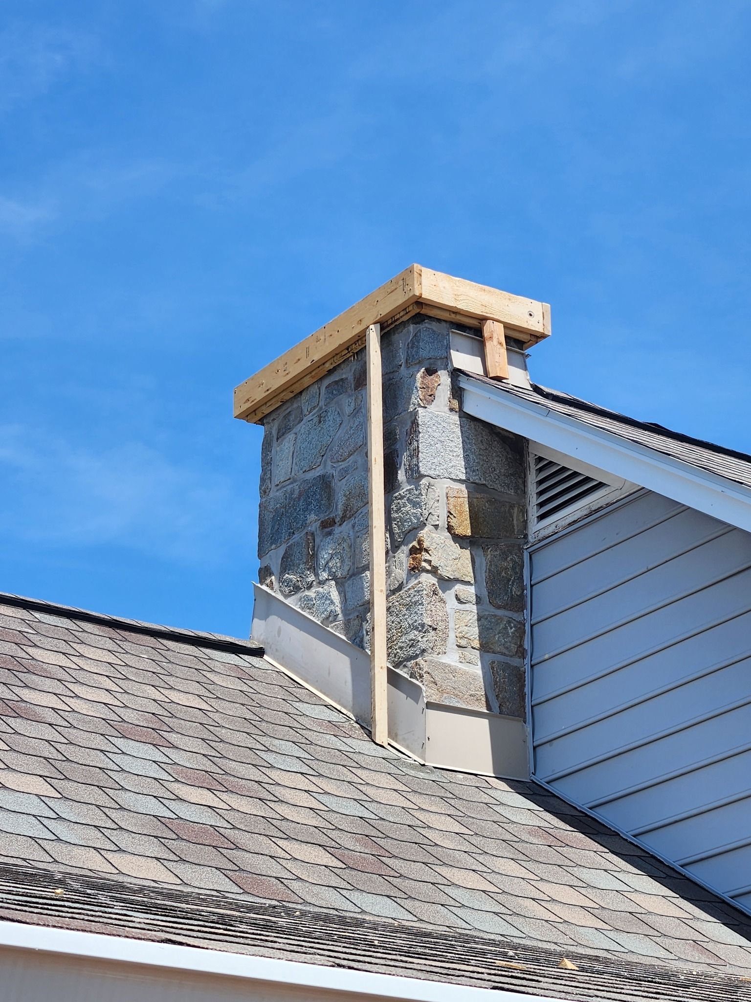 Stone chimney on a shingled roof under a blue sky, with wood framing around the top.