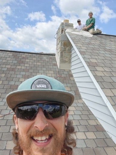 Man smiles into camera with two workers on a rooftop near a chimney, blue sky background.