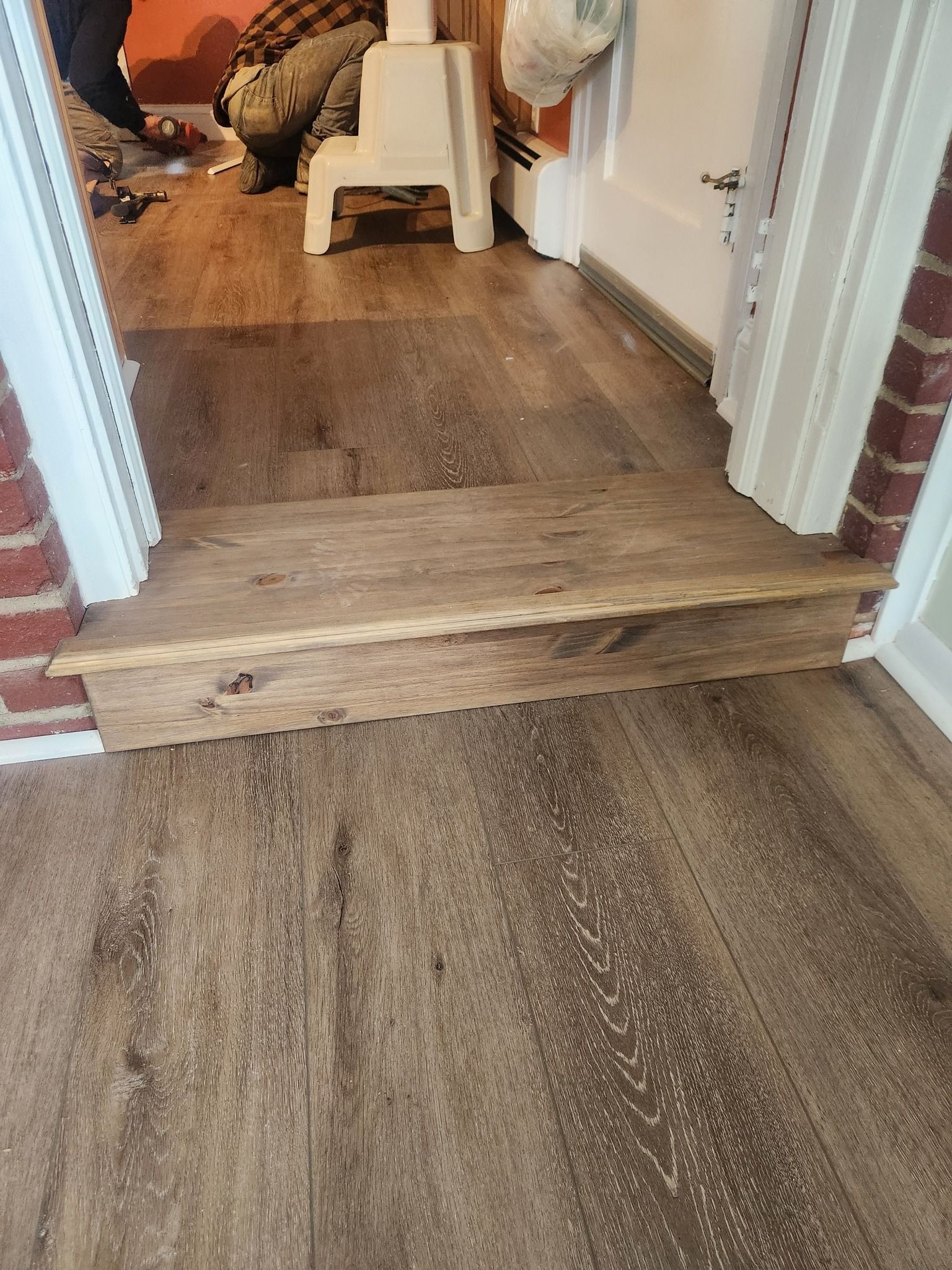 Wooden step in a doorway with wood-look flooring. White trim on the door frame and red brick on one side.