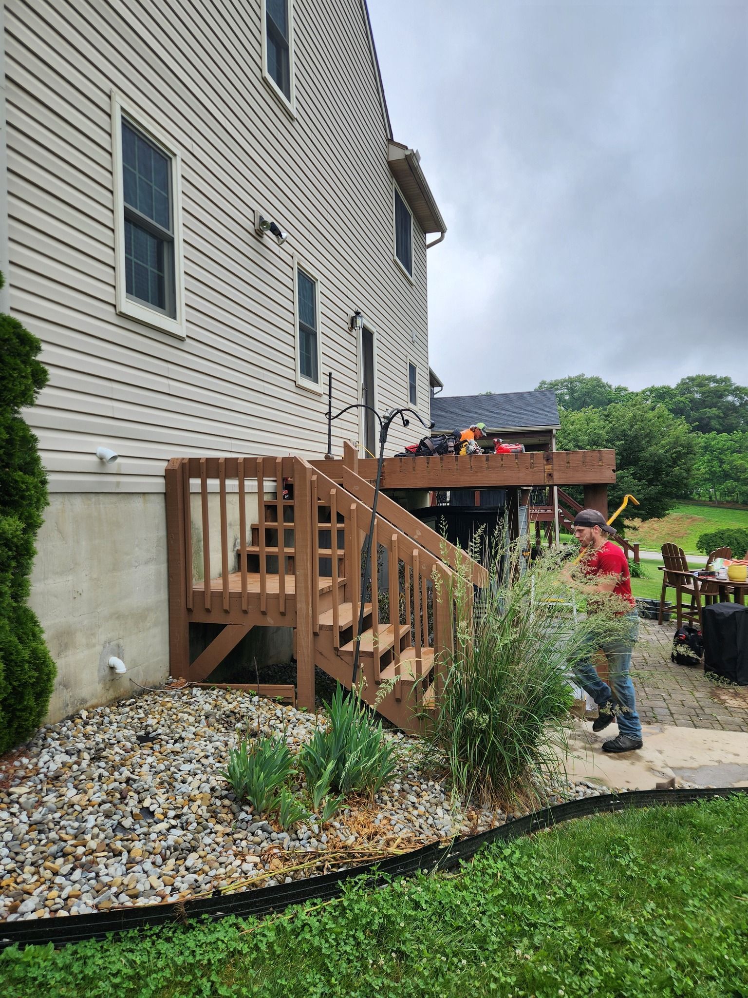 Brown wooden deck stairs leading to a deck. A person stands near plants. Beige house exterior.