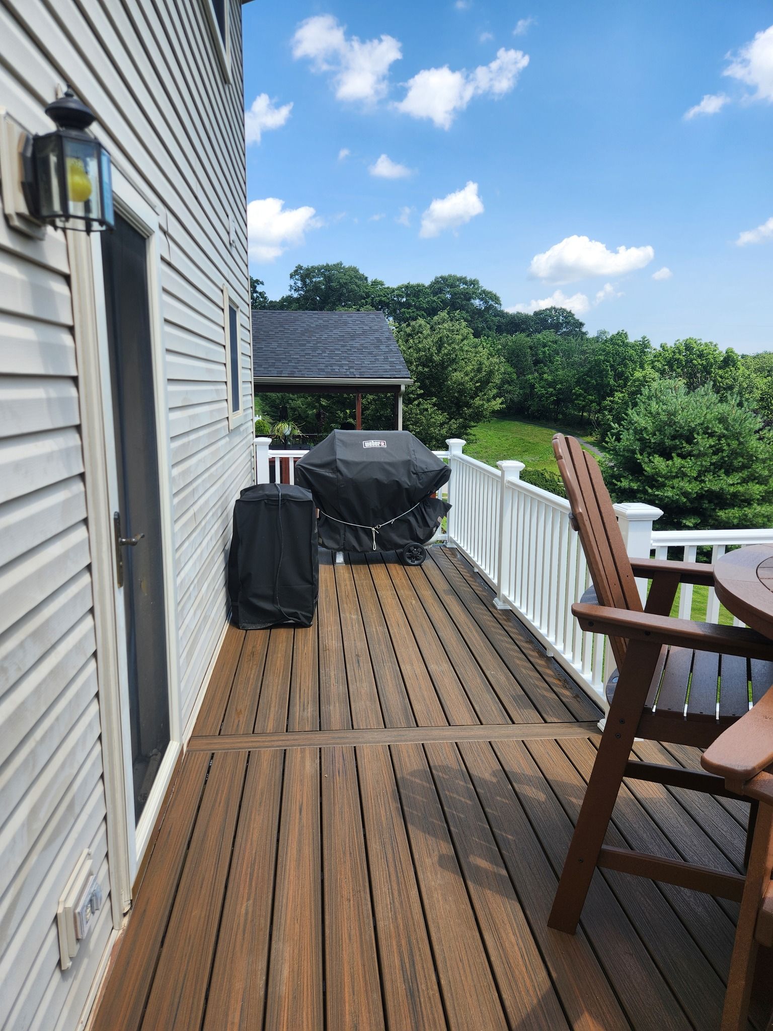 Deck with a grill and a chair, overlooking trees and a blue sky.
