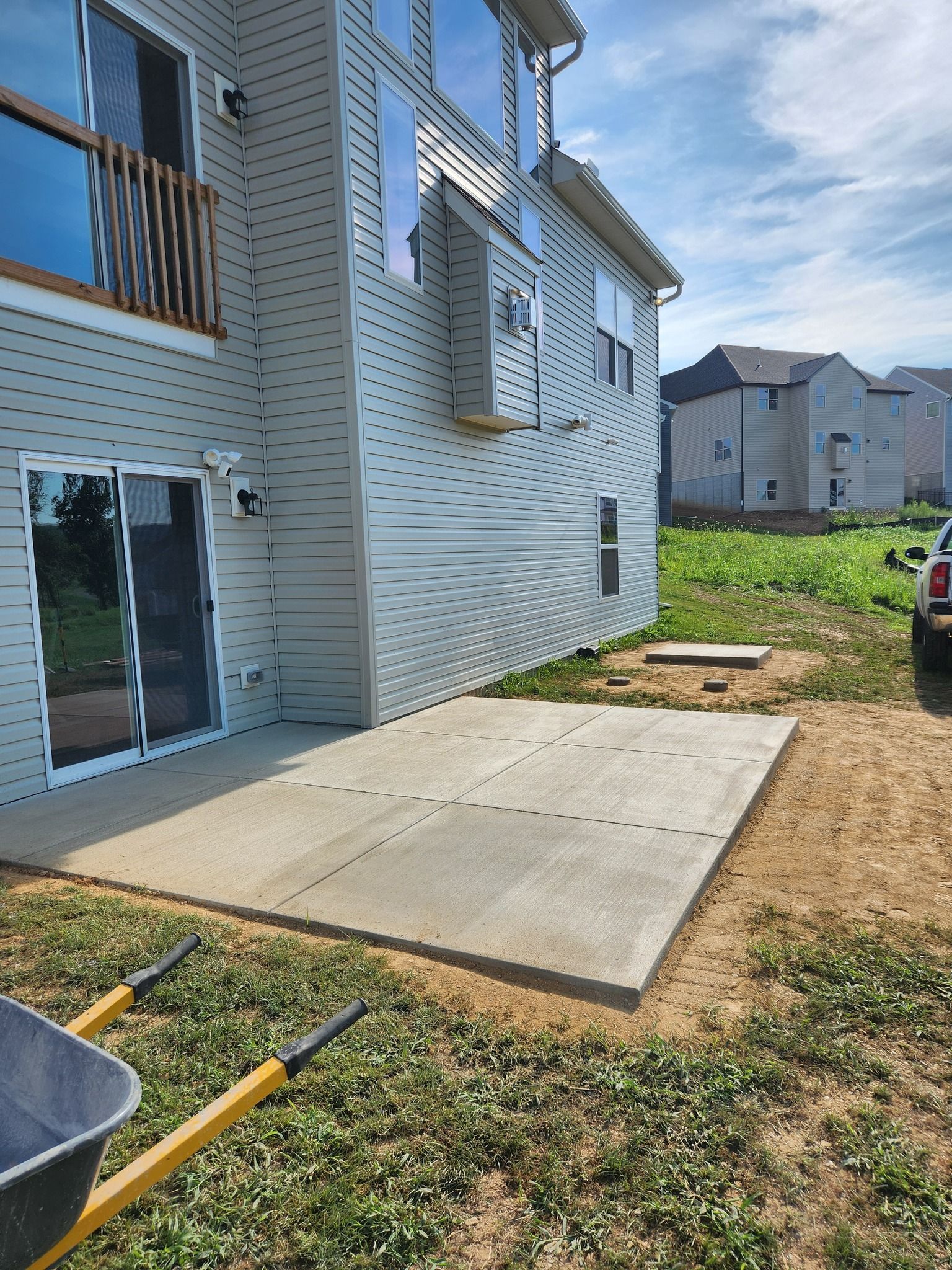 Back of a two-story house with a newly poured concrete patio. A wheelbarrow sits nearby on the grass.
