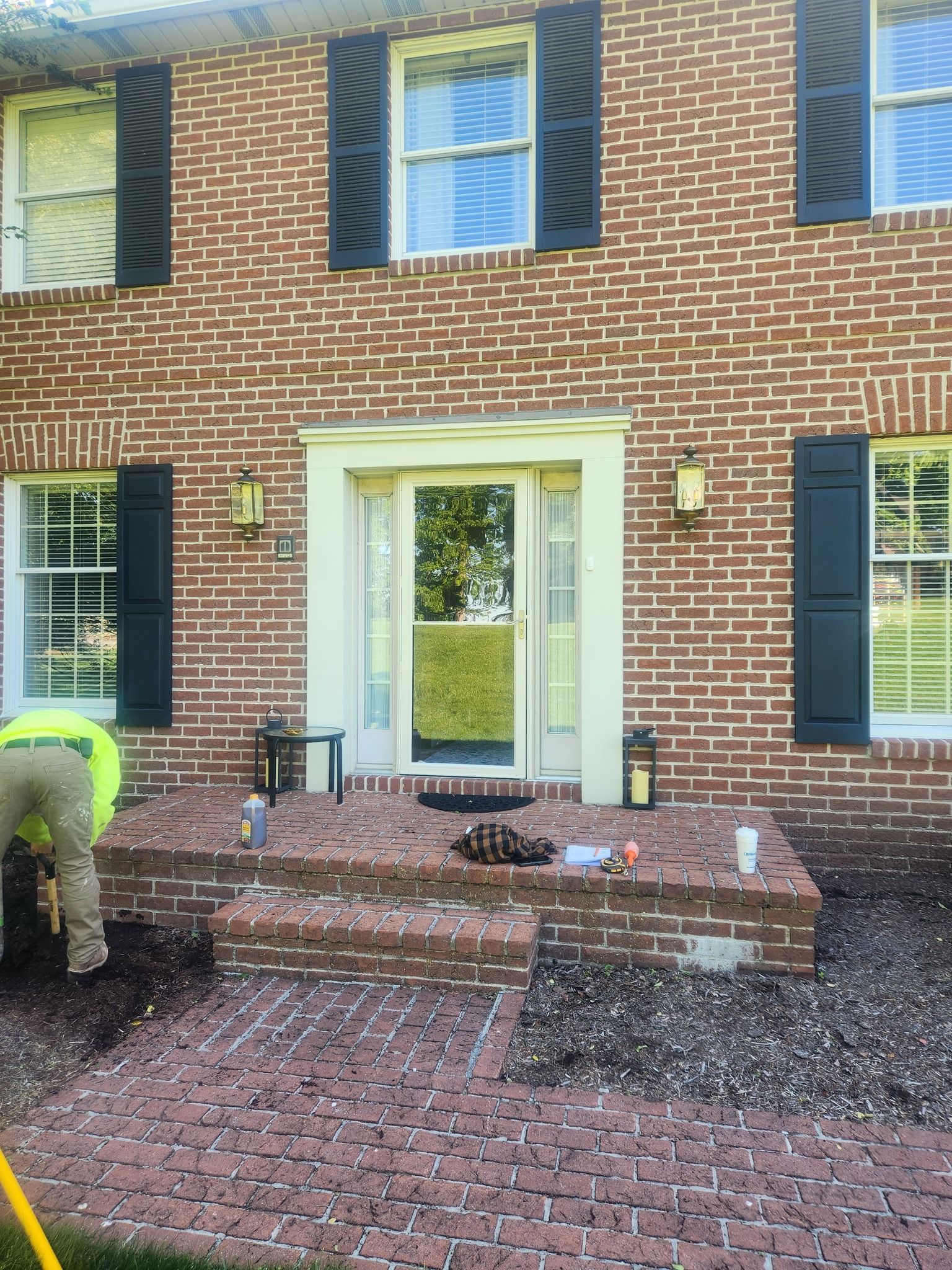 Brick home with person working on brick walkway near front door. Black shutters, beige door frame.