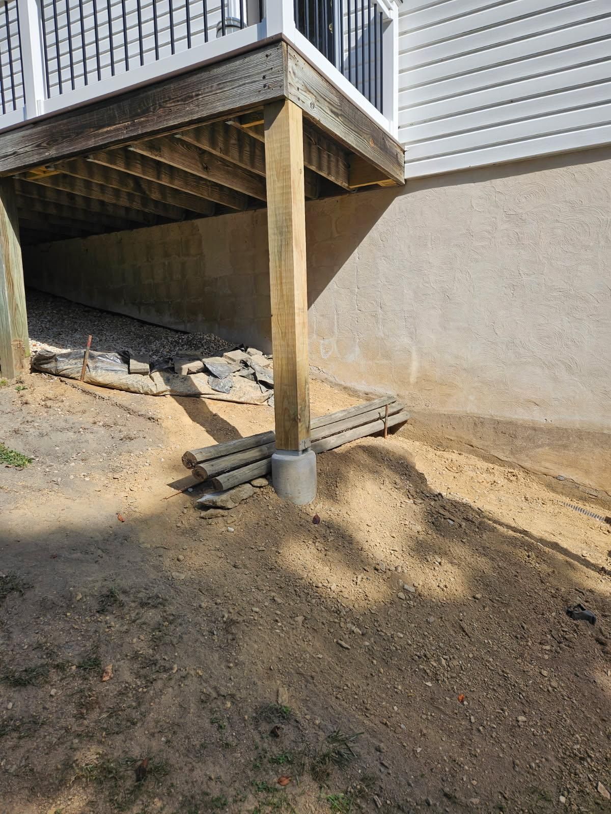 View of deck supported by wooden posts on a sloped, dirt yard with a retaining wall.