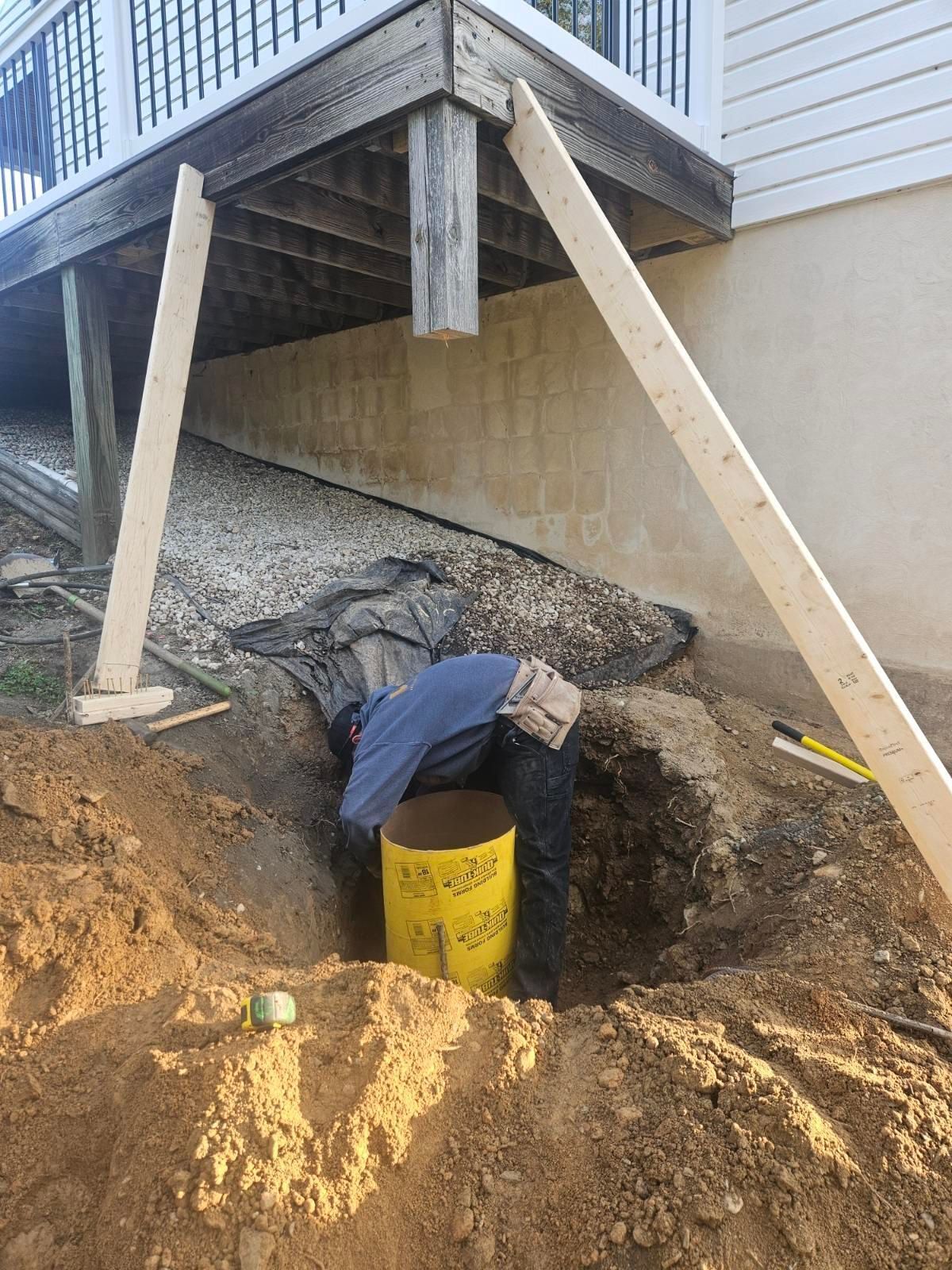Person working on construction project under a deck, digging in a hole with a yellow cylinder visible.