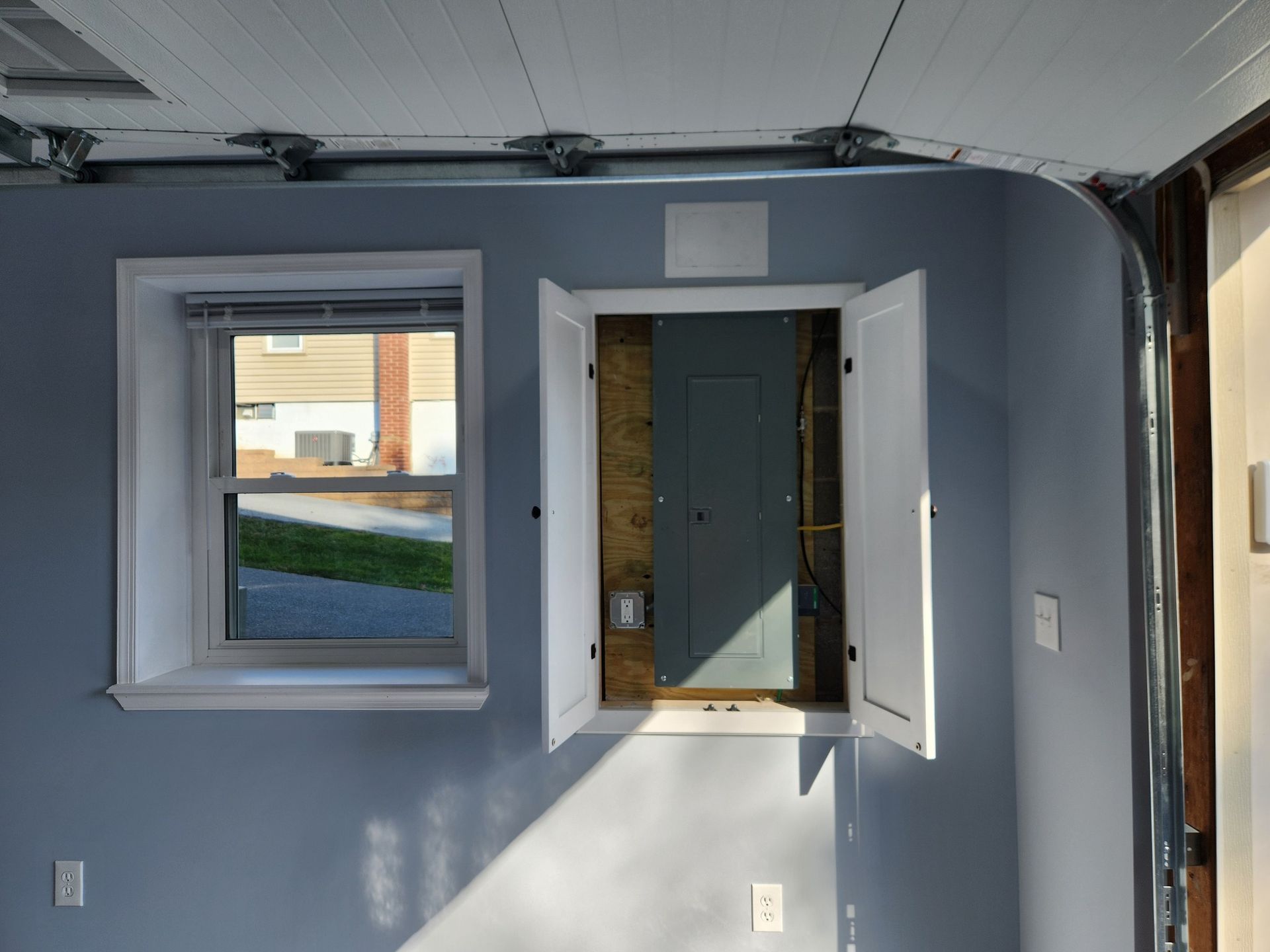 Blue wall with a window and electrical panel with open doors, in a garage.