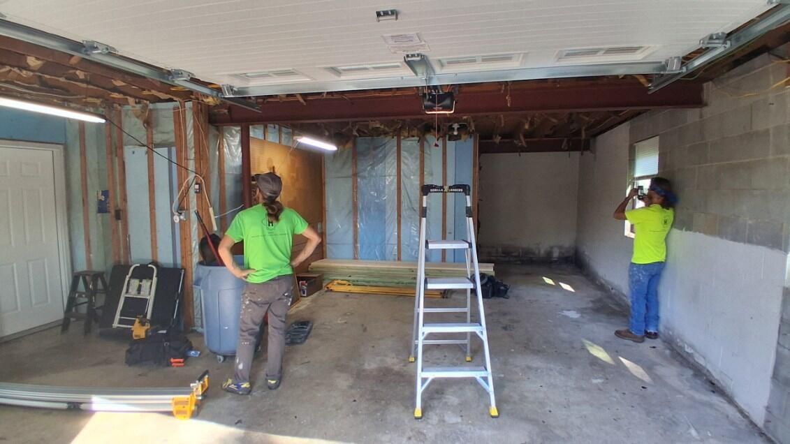 Two people stand in a gutted garage with exposed framing. One person is posing, and the other is taking a picture.