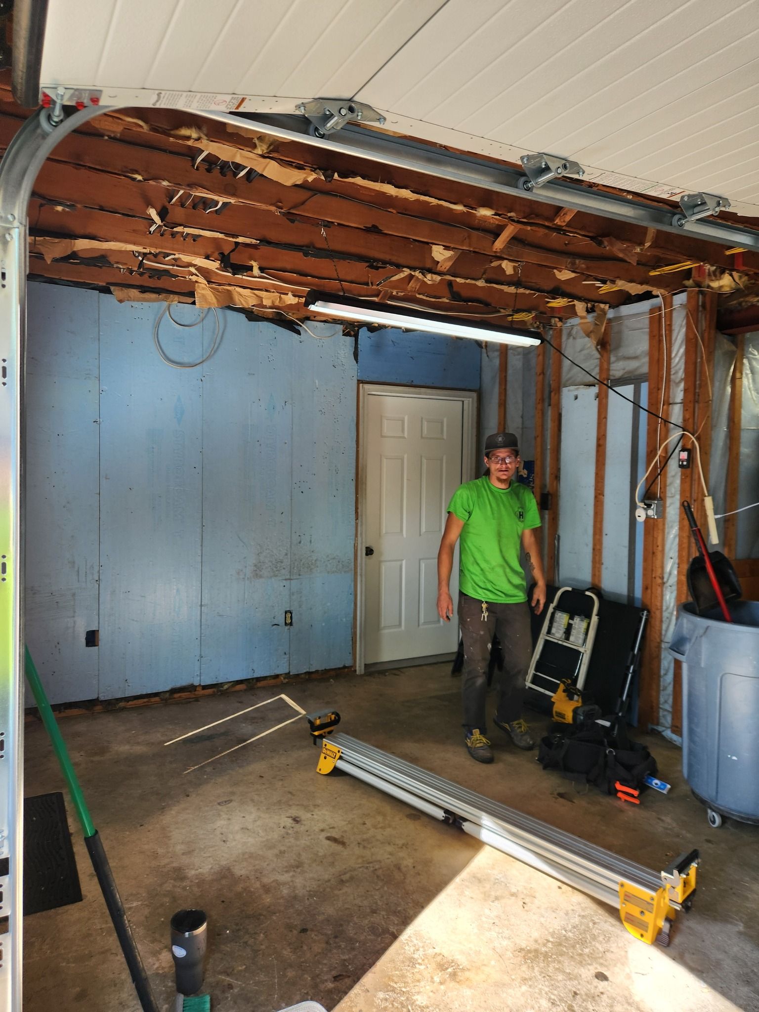 Man standing in a garage with damaged ceiling and open door, tools and debris visible.