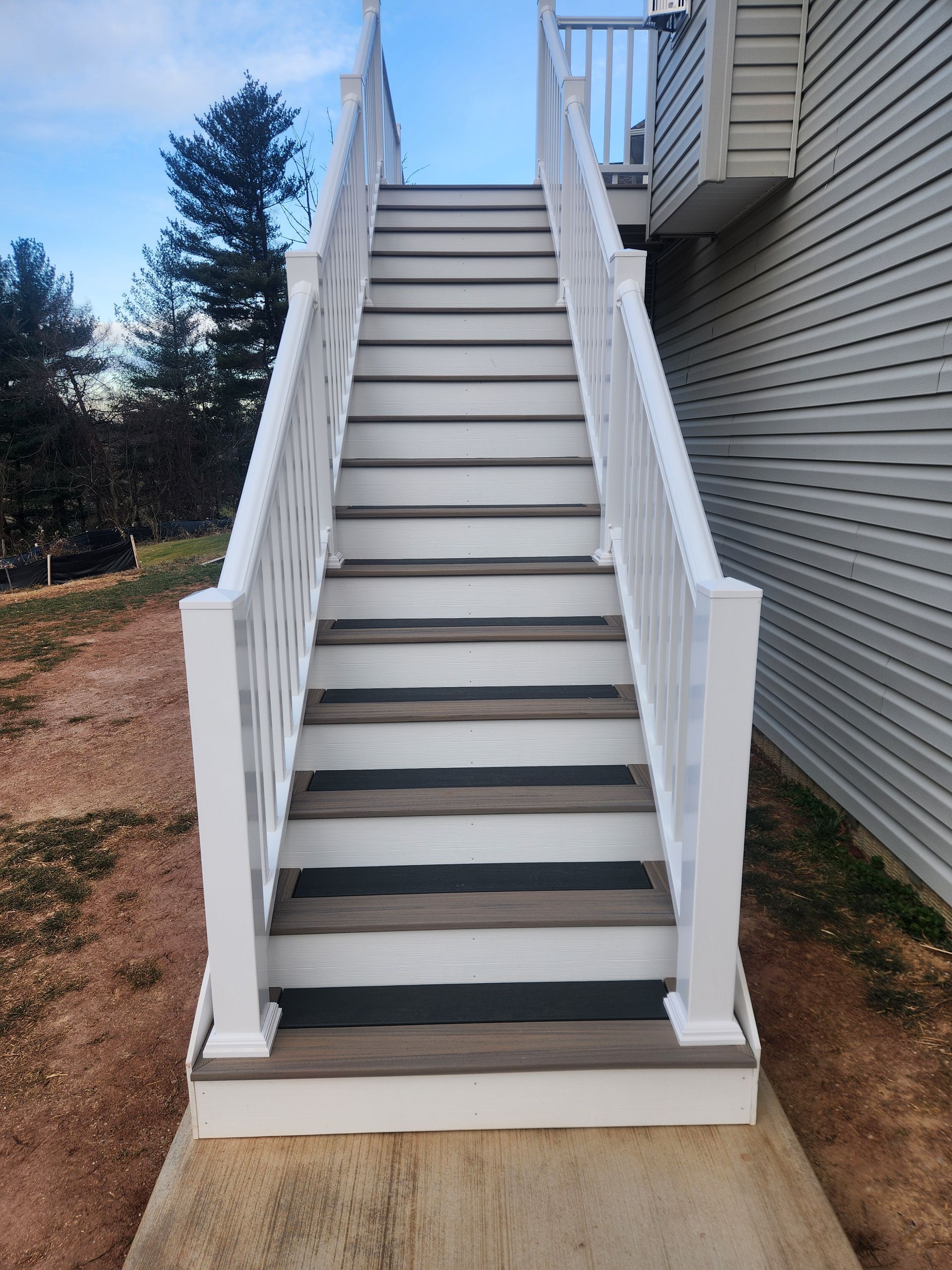Outdoor wooden staircase with white railings, leading up to a house with light grey siding on a dirt ground.