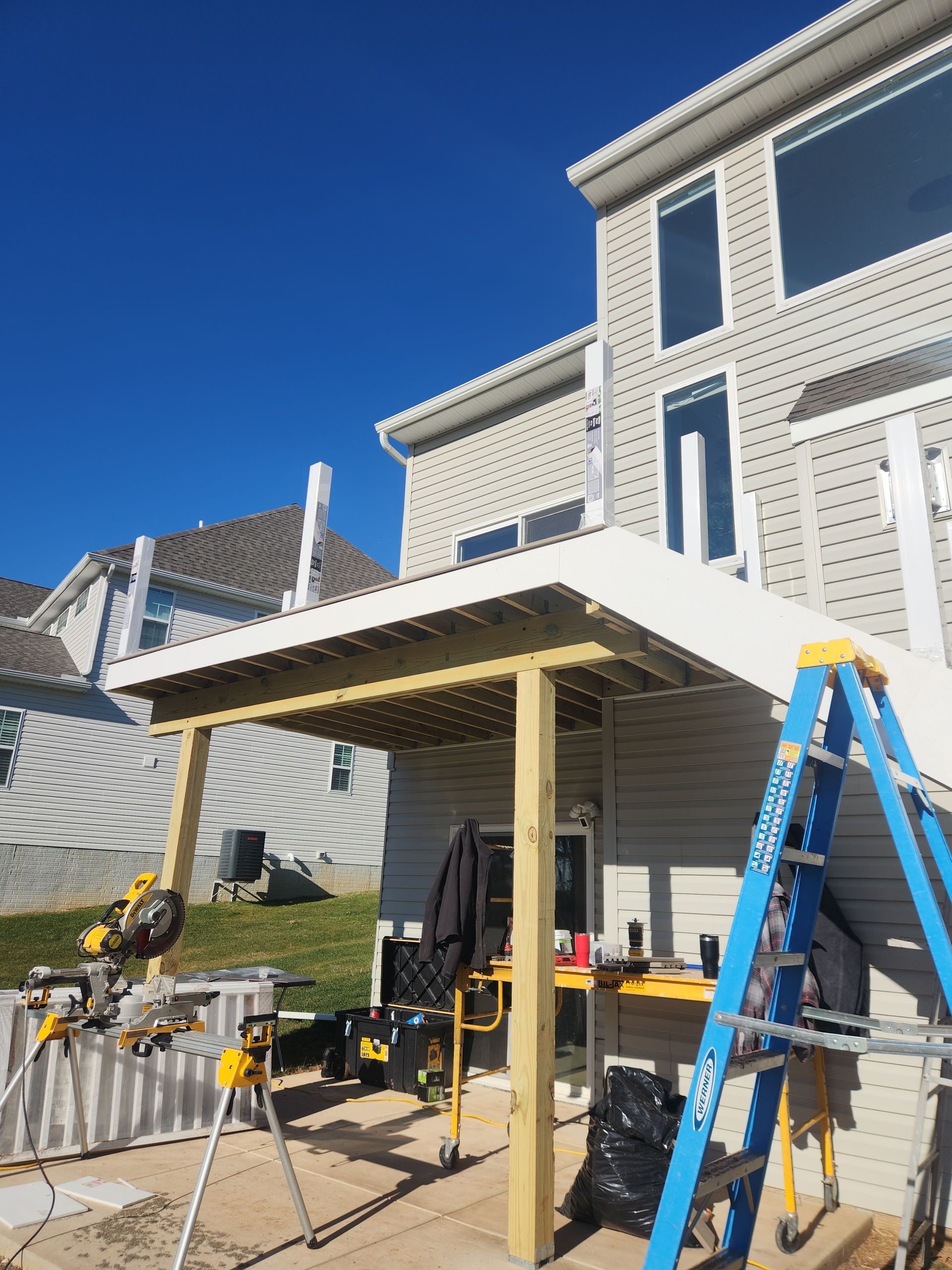 A construction site showing a house with a newly framed deck, wooden support posts, a miter saw, and a blue ladder.