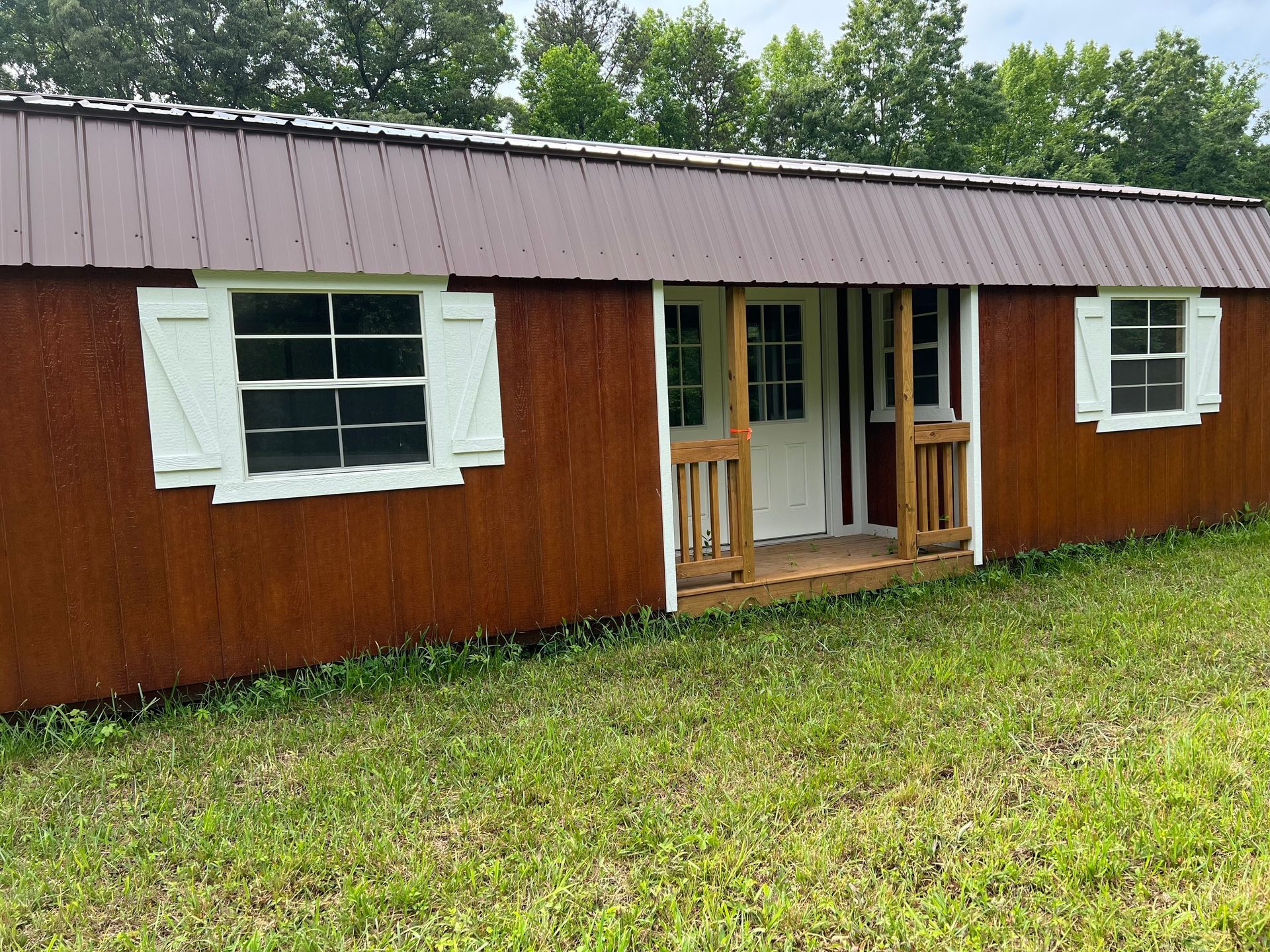 Brown shed with a brown metal roof, white window frames and shutters, and a small wooden porch, set in a grassy area.