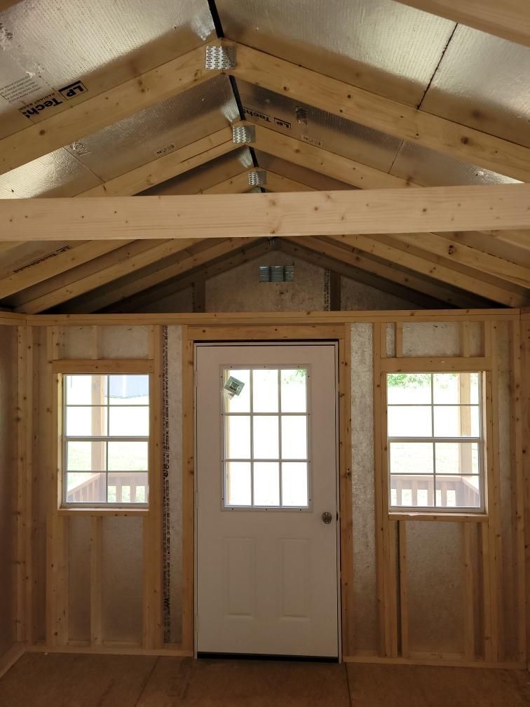 Interior view of a wooden shed with a door, two windows, and exposed rafters. The walls and floor are unfinished.