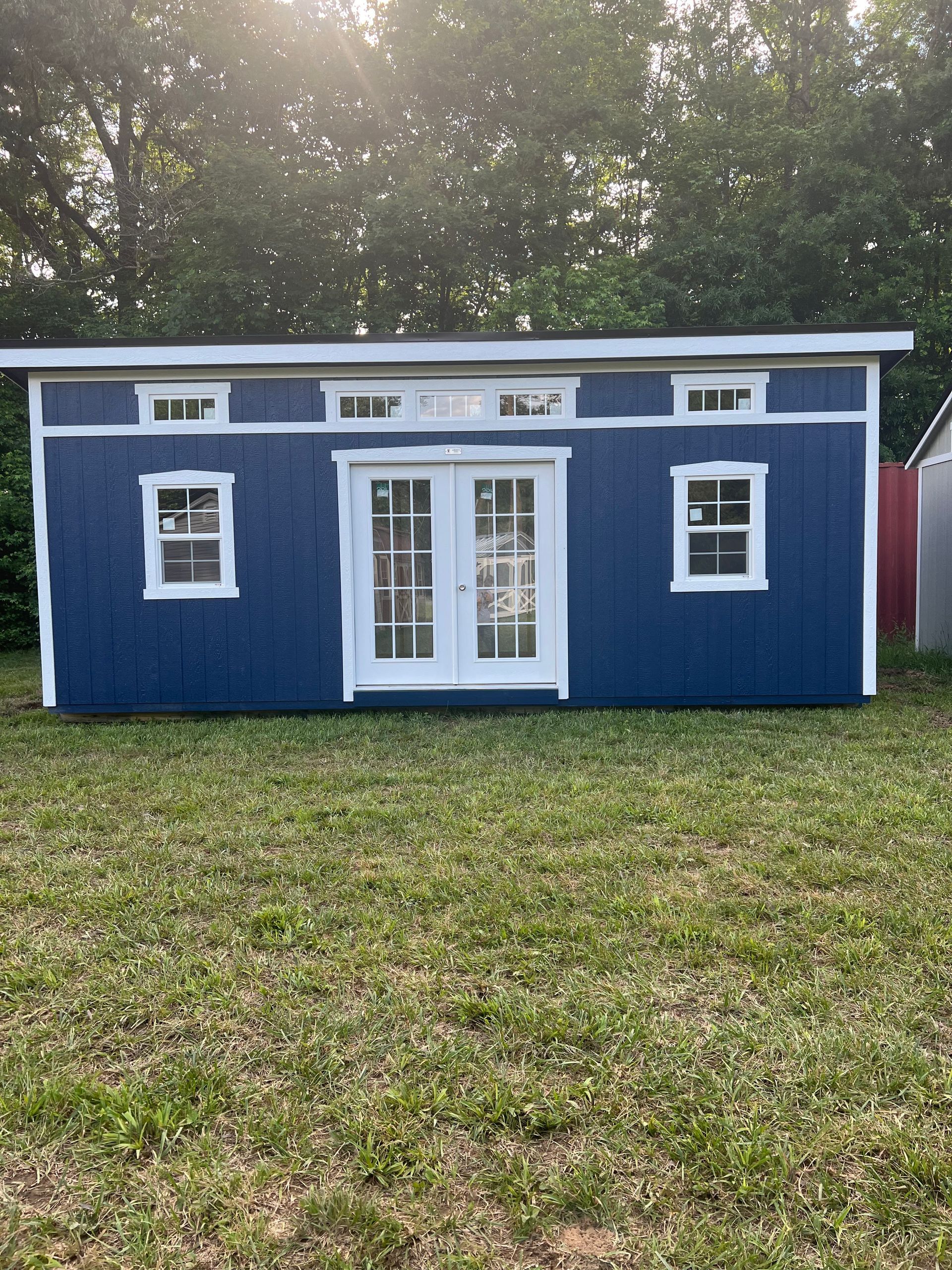 Blue shed with white trim, French doors, and windows, sitting on green grass. Sunlight shines from above.