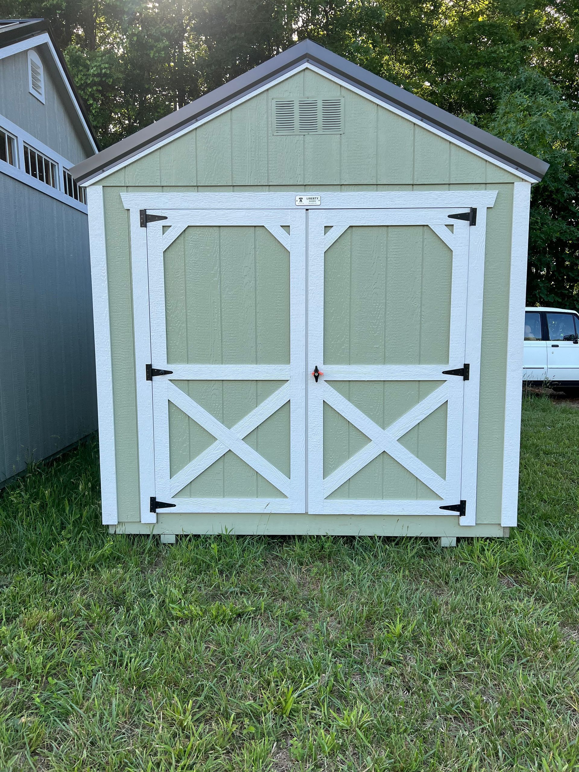 A green shed with white trim and double doors stands on grass, with a brown roof and a small window.