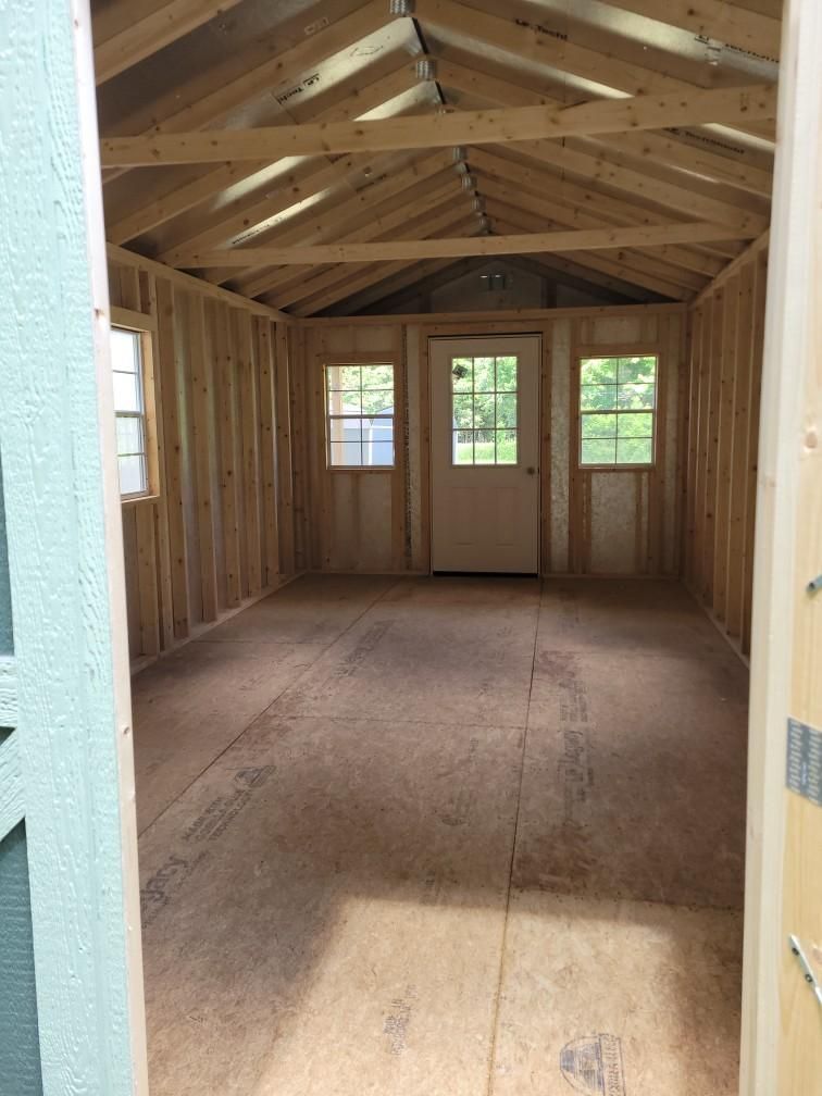 Interior of a wooden shed with a door and windows. The room is empty with a plywood floor and unfinished wooden walls.