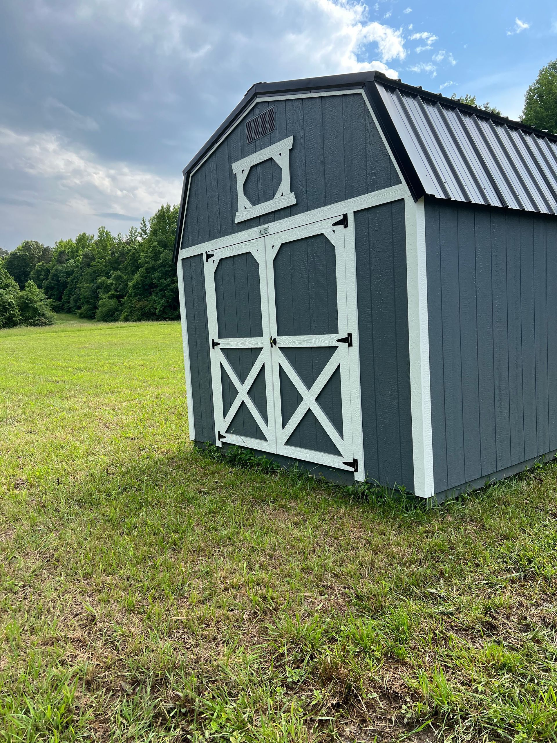 A small, gray barn-style shed with white trim and a black metal roof stands in a grassy field under a cloudy sky.