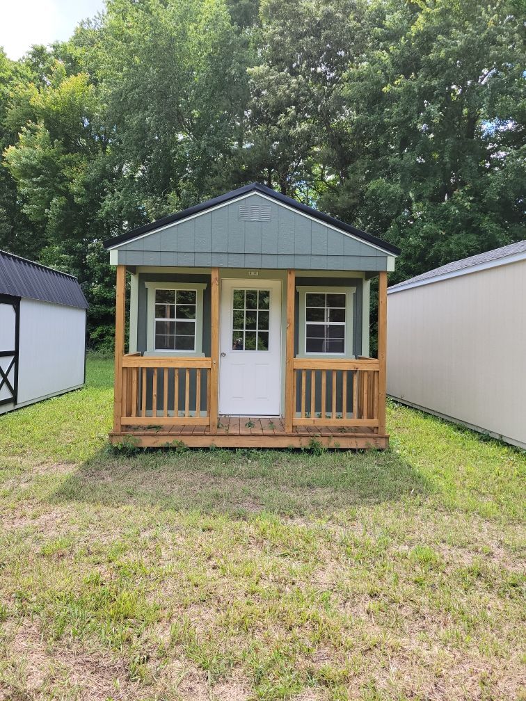 Small teal shed with a porch and white door, set on grass with other sheds nearby.