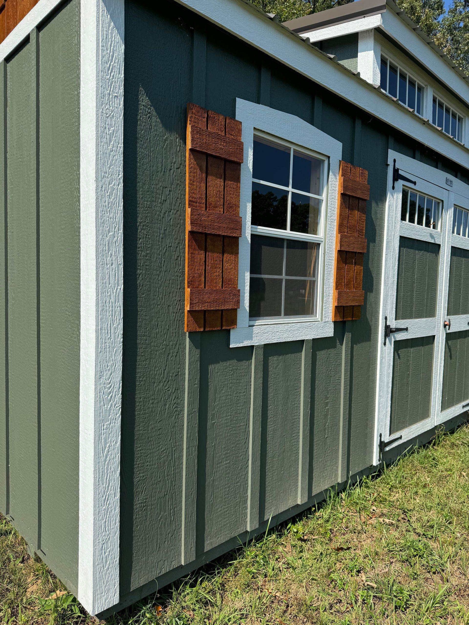Green shed with white trim, brown shutters and a small window; building is in a grassy area.