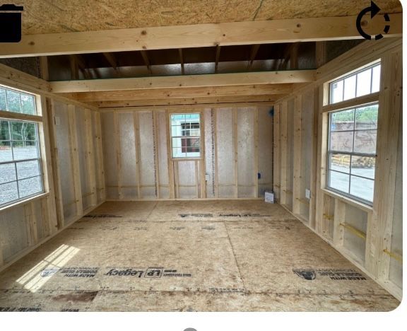 Interior view of a wooden shed under construction, with windows and exposed framing. The floor and walls are unfinished.