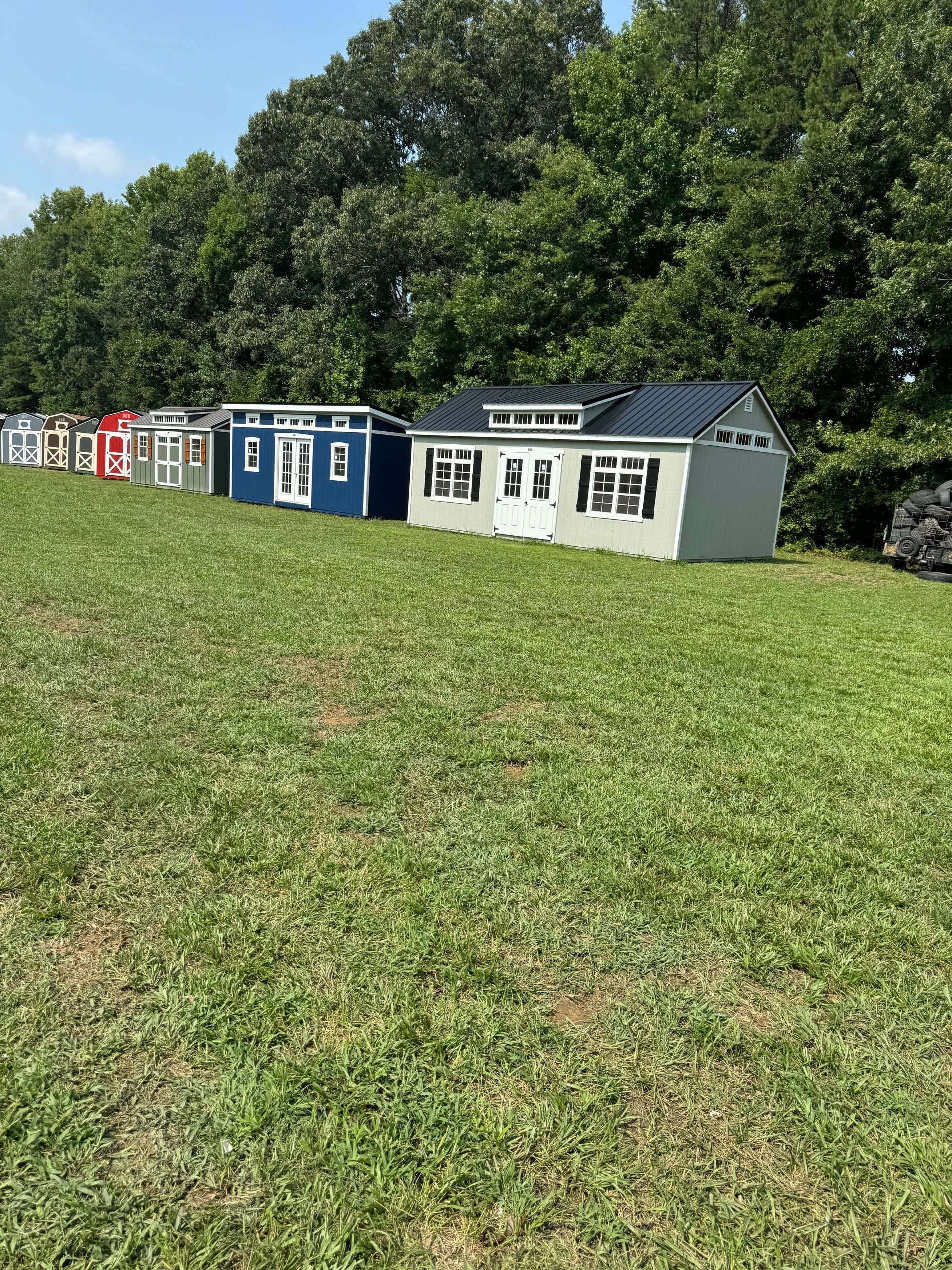 Row of colorful sheds lined up on a grassy field, set against a backdrop of trees under a bright sky.