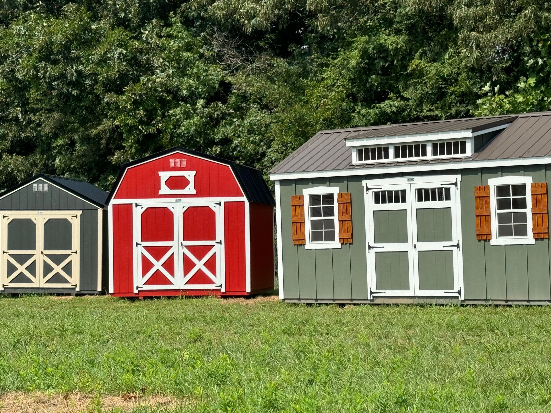 Three colorful garden sheds on grass. One gray, one red barn style, and one green with a brown roof.