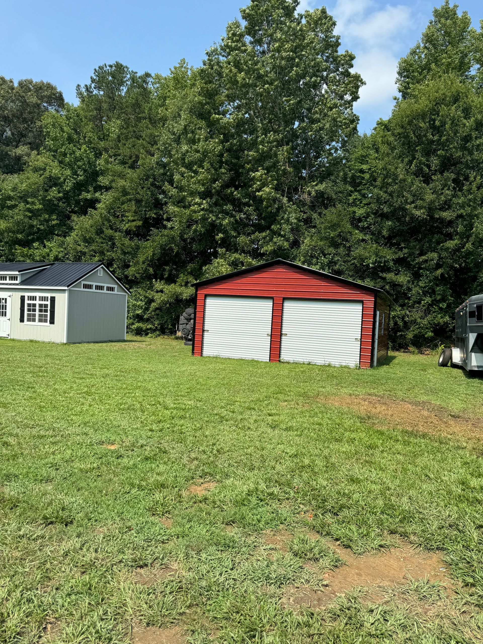 Red garage with white garage doors sits in a grassy yard with two other buildings and trees in the background.