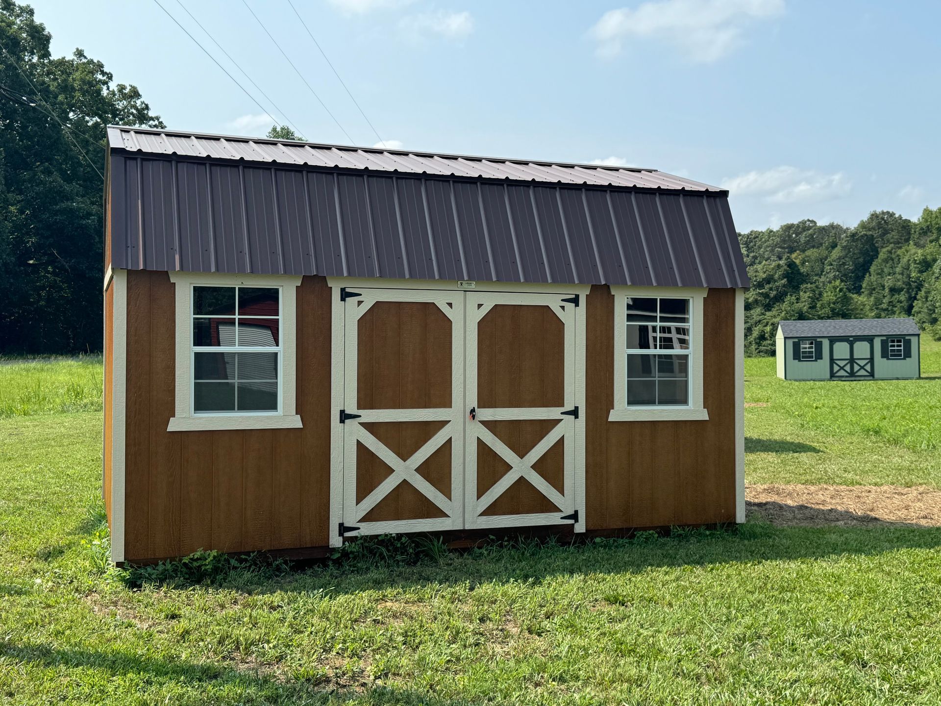 Brown storage shed with white trim and a brown metal roof. In a grassy field, another shed is visible in the background.