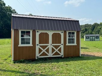 Brown wooden shed with white trim and a brown metal roof, set in a grassy field on a sunny day.