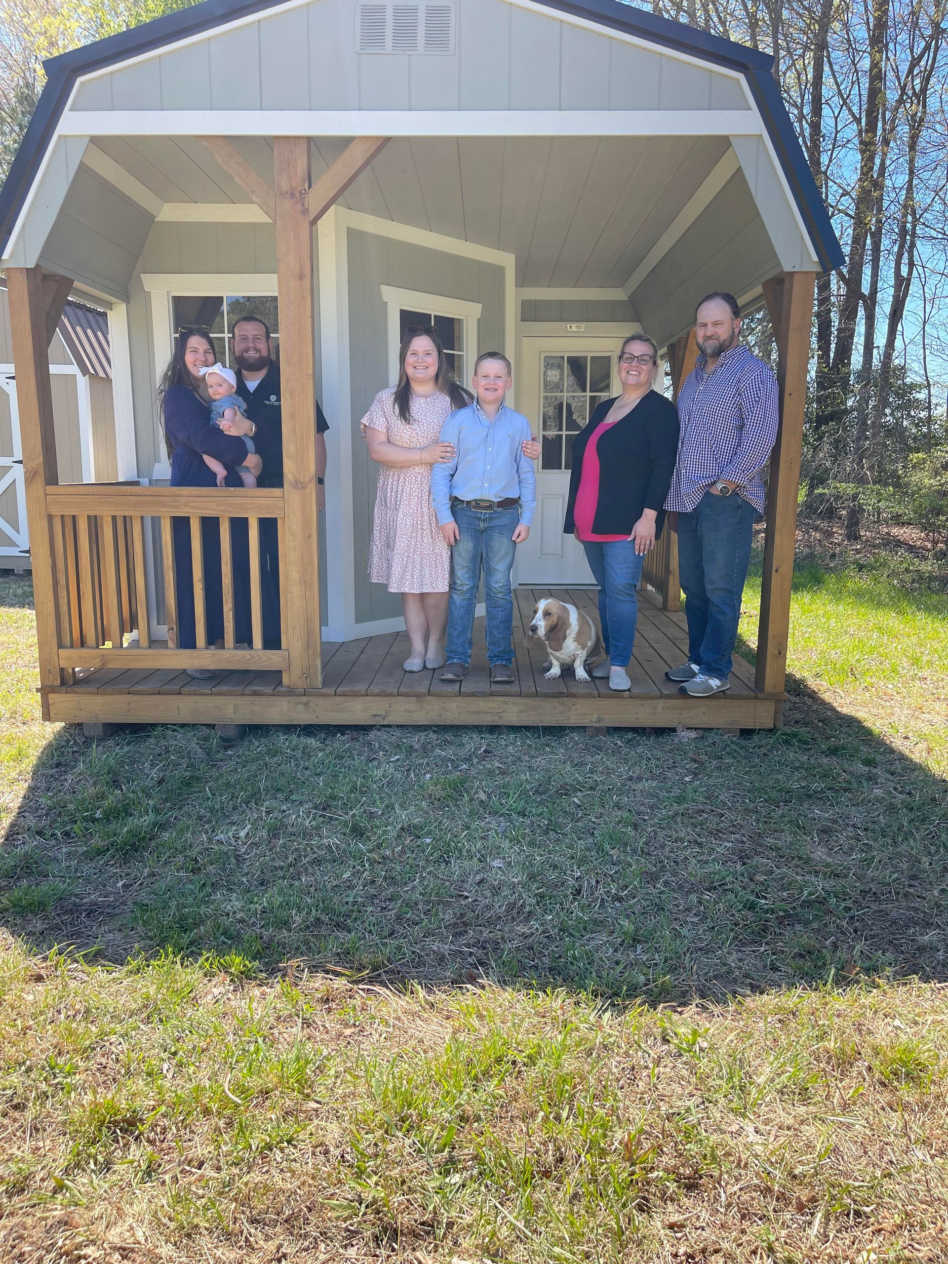 Family posing on the porch of a small, light-green shed-like structure with a dog.