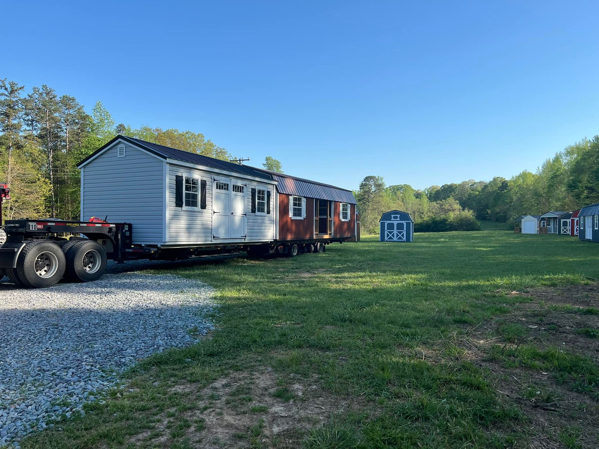 A tiny house, light blue with brown accents, is being transported on a large trailer on a grassy field.