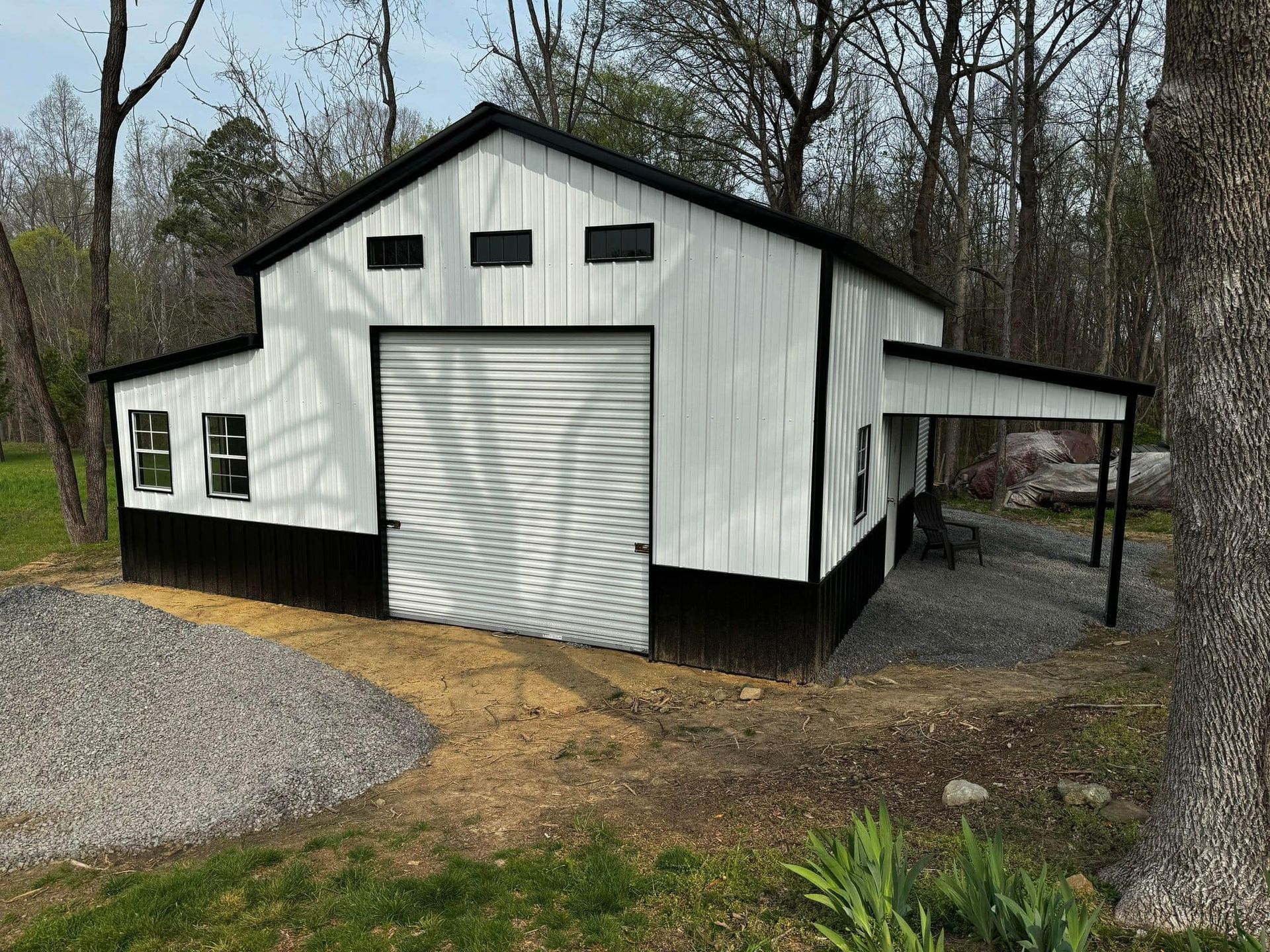 White and black barn with a large garage door and a small overhang. It sits on a gravel driveway surrounded by trees.