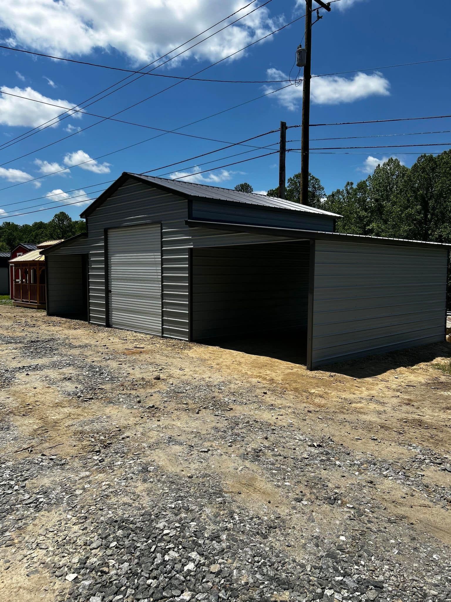 Gray metal shed with a roll-up door, surrounded by gravel on a sunny day. Power lines and trees are in the background.