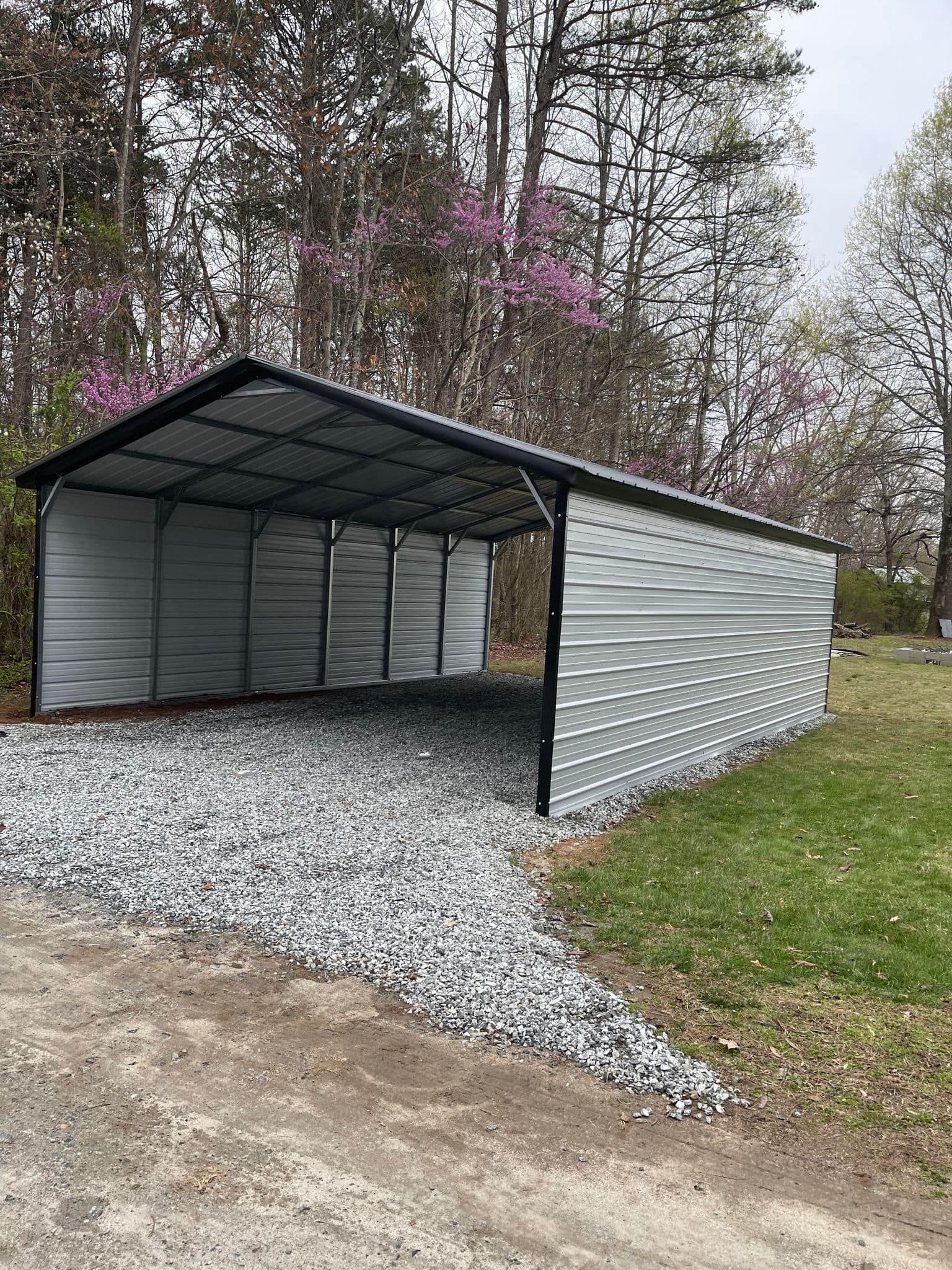 Metal carport shelter with a gravel driveway in a grassy setting with trees. The roof is black, and the structure is silver.