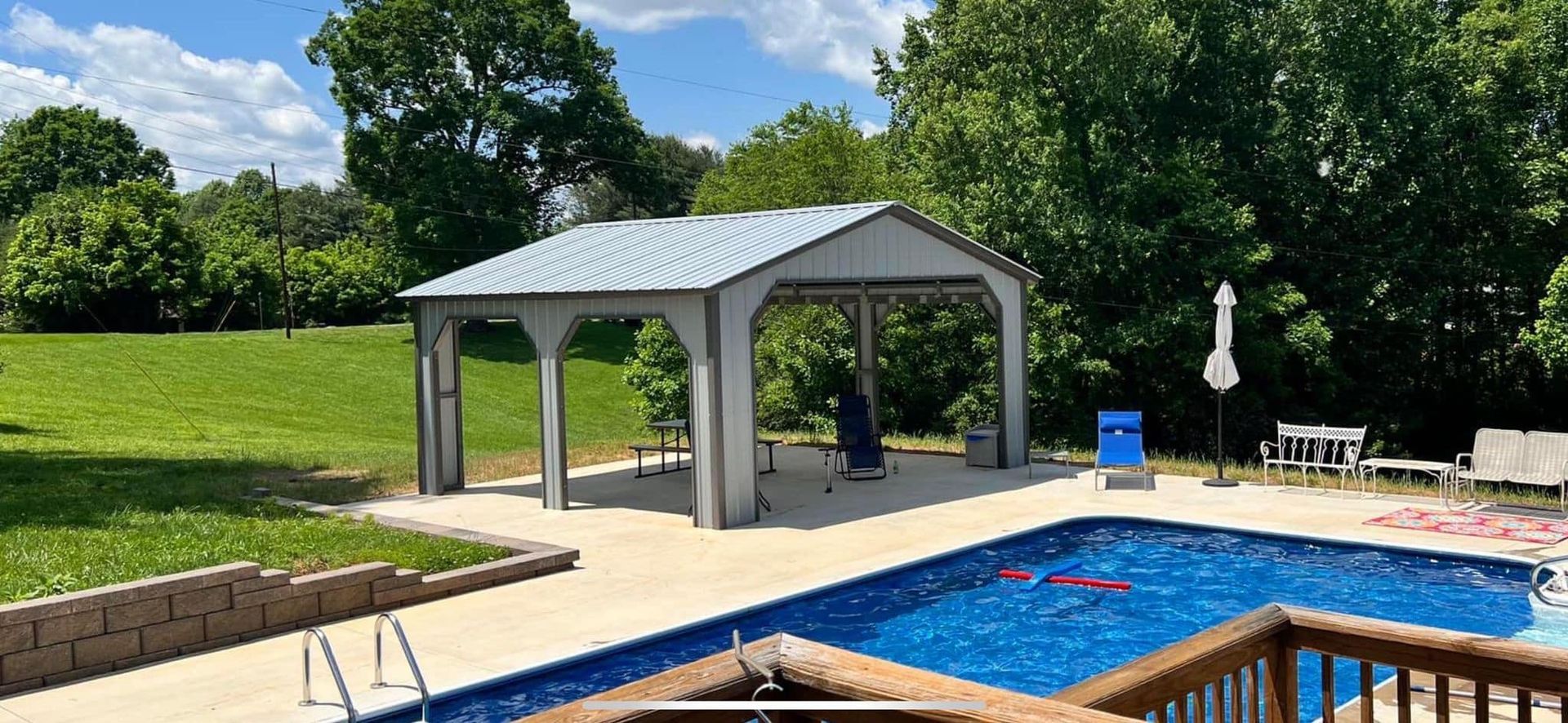 Backyard scene with a swimming pool, patio area, gazebo, and green lawn. Trees and a bright blue sky are in the background.