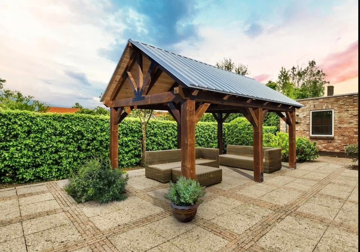 Wooden gazebo with metal roof, straw-bale seating, on a paved patio, against a backdrop of a green hedge and a colorful sky.