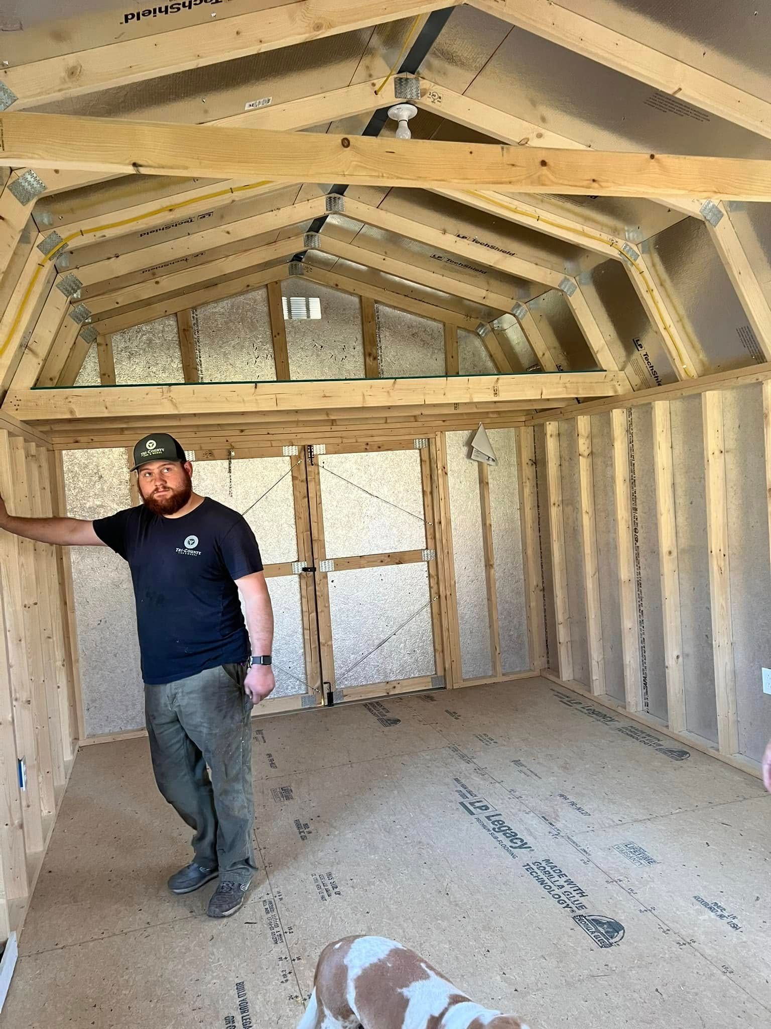 A man stands inside a wooden shed under construction. He wears a dark shirt, work pants, and a cap