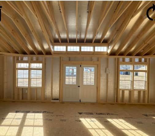 Interior view of a building under construction with wooden framing and windows. The room is empty with sunlight streaming in.