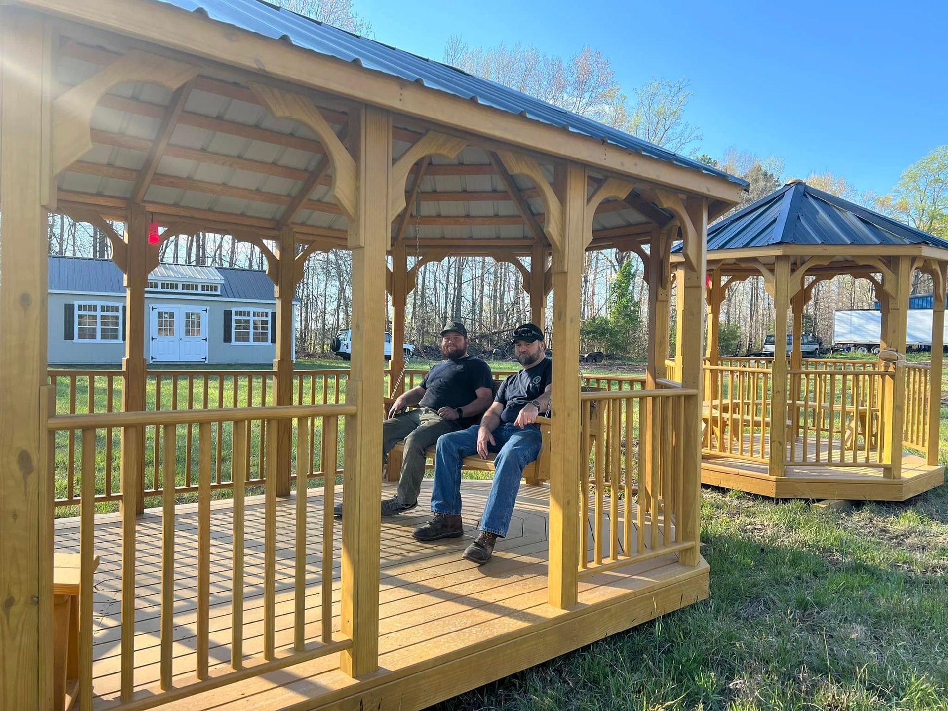 Two men sit in a light-colored gazebo with a metal roof. Another gazebo is visible behind them.