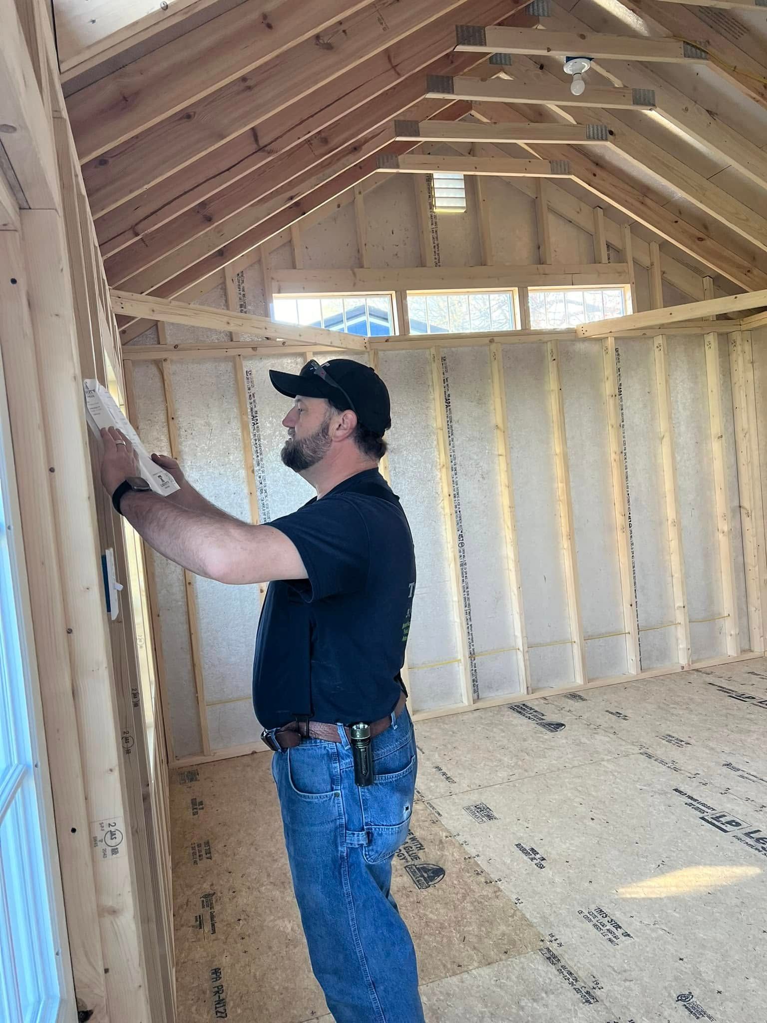 A man in jeans and a black shirt measures a wooden wall inside a building under construction.