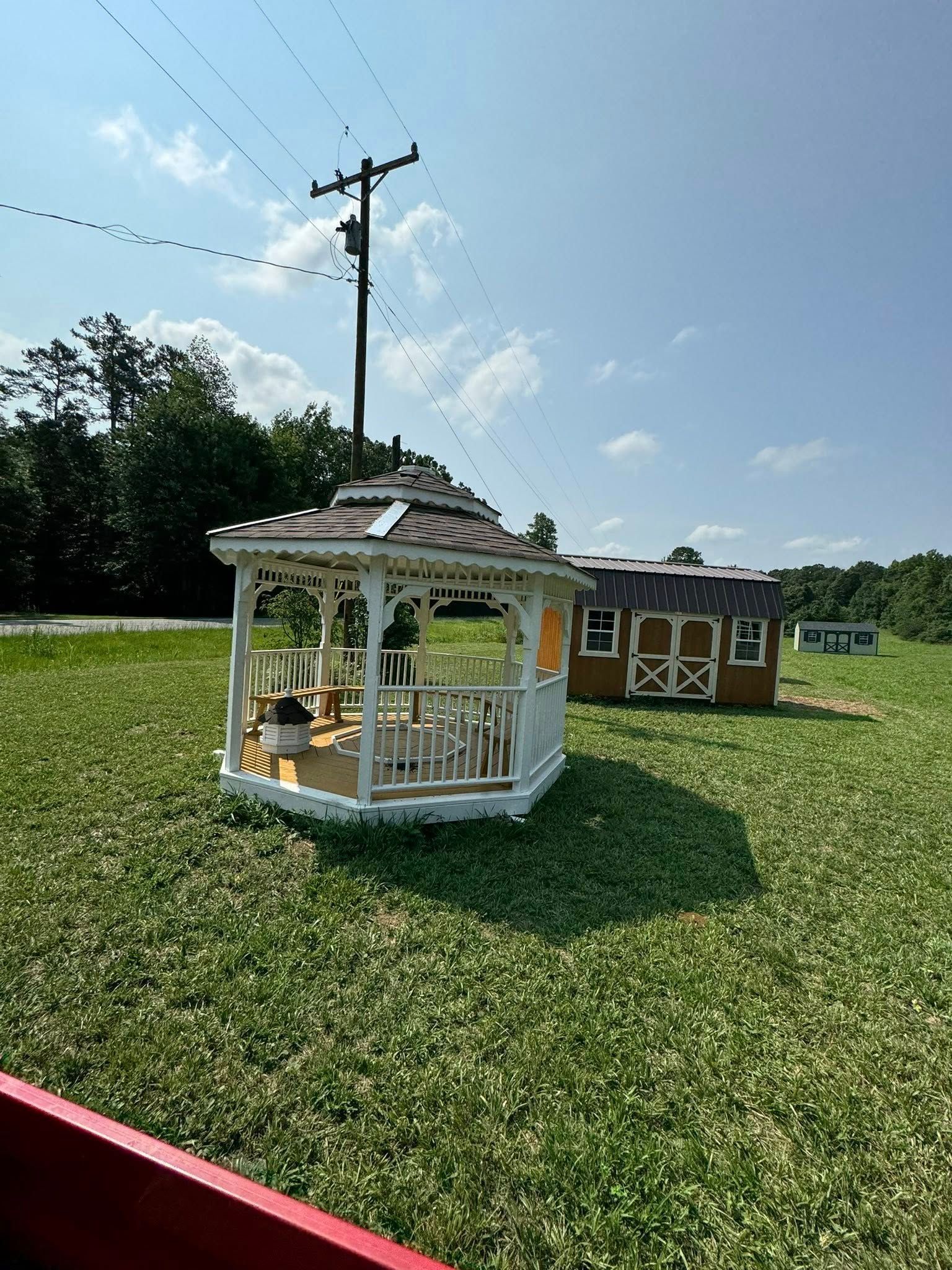A white gazebo and brown shed on a grassy field under a blue sky, with a utility pole in the background.