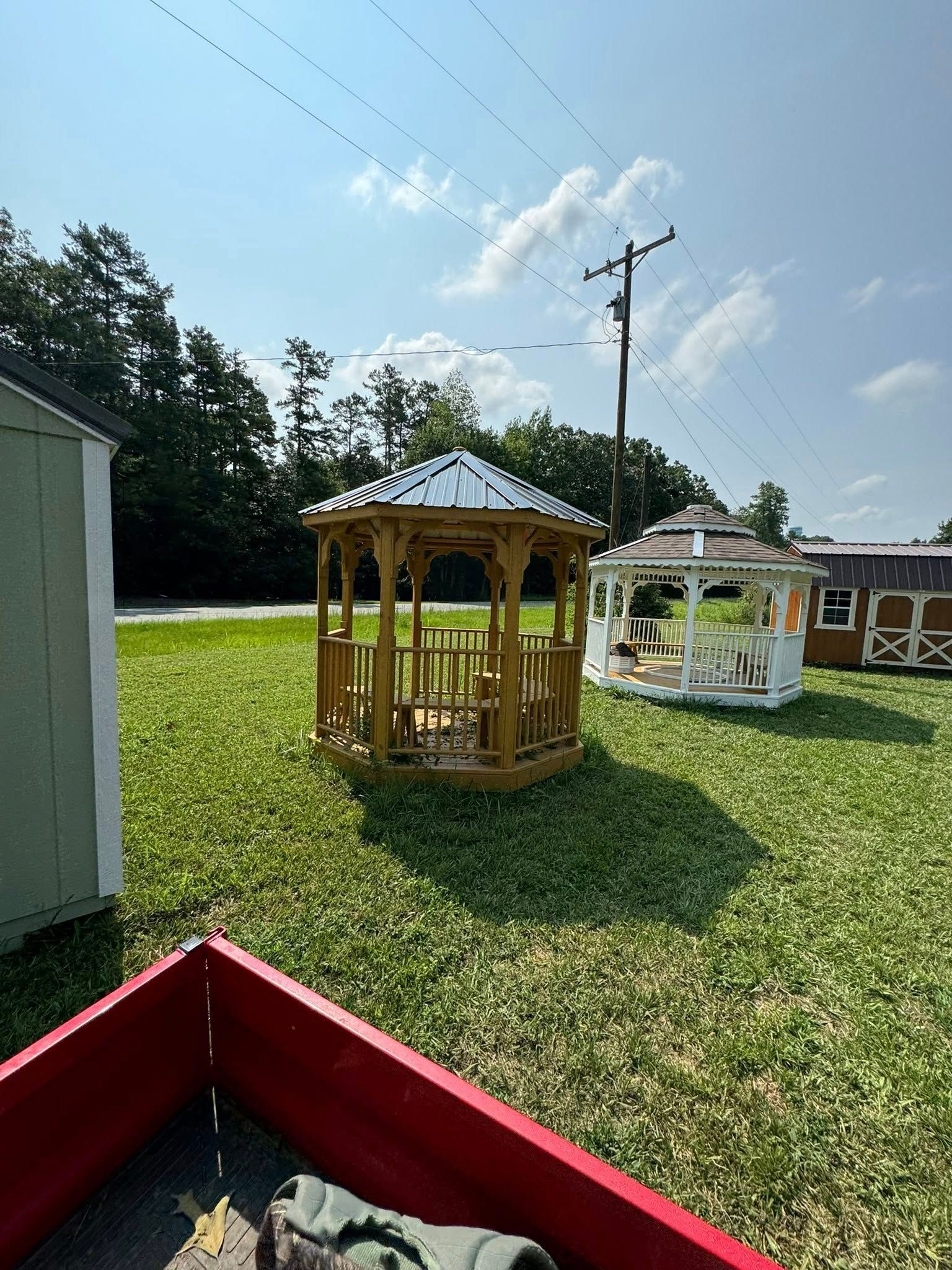 A wooden gazebo with a metal roof on a grassy lawn, with other structures and a clear sky.