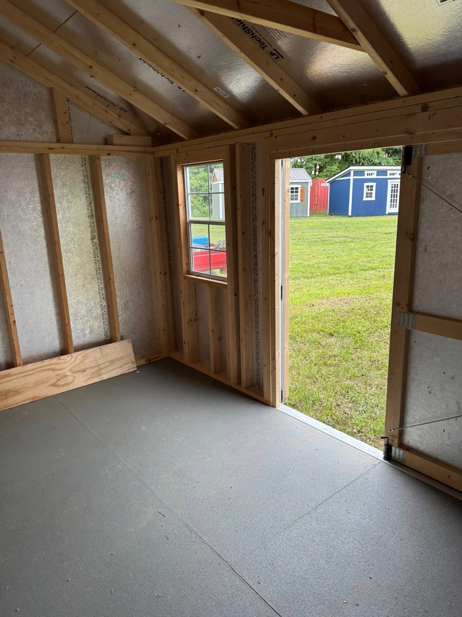 Interior view of an unfinished shed with an open doorway, window, and gray flooring.