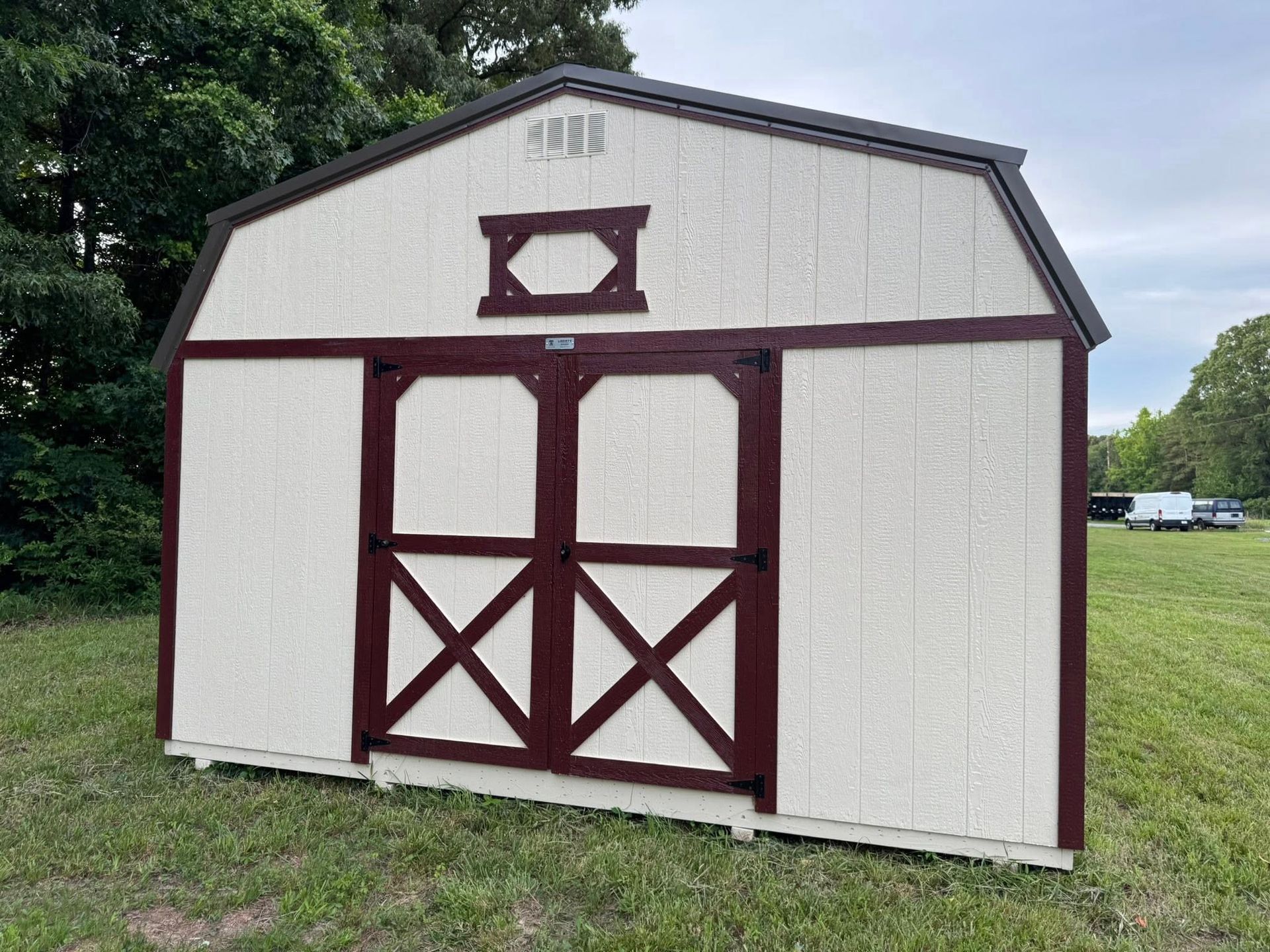 White and burgundy barn-style shed with double doors in a grassy field. The roof is brown.