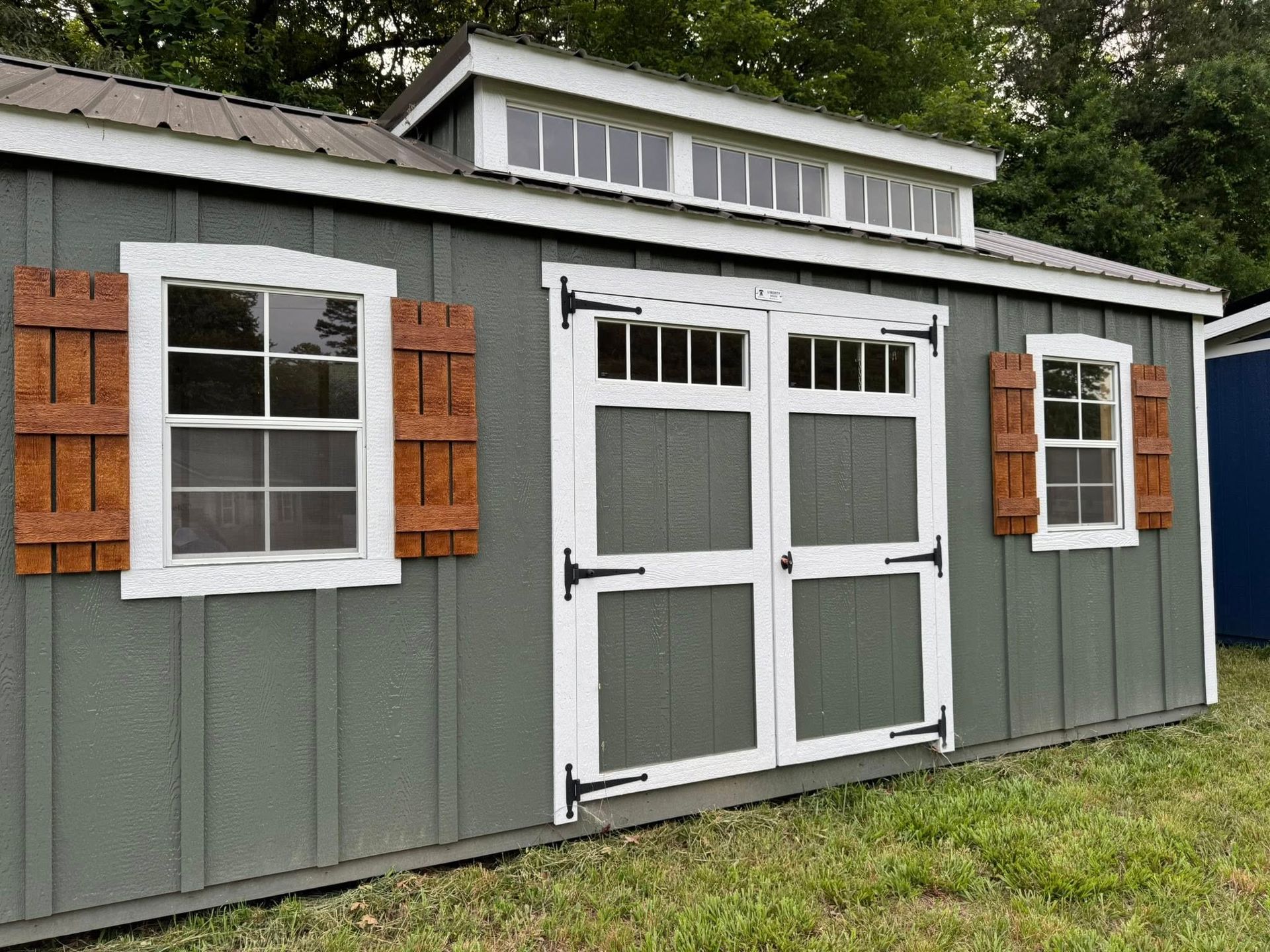 A green shed with white-framed windows and doors, brown shutters, and a row of windows along the roofline.