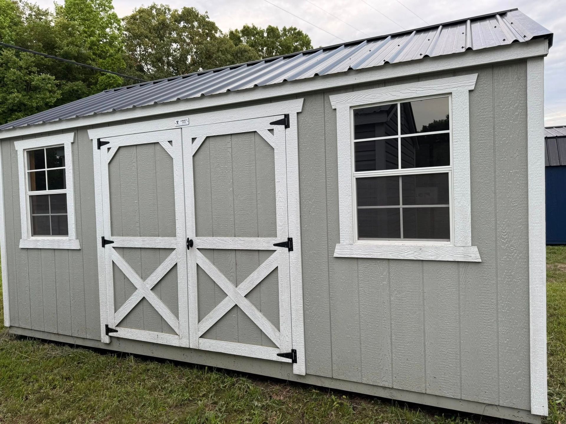 Gray shed with white trim, door, and windows. Metal roof.  Set outdoors on grass.
