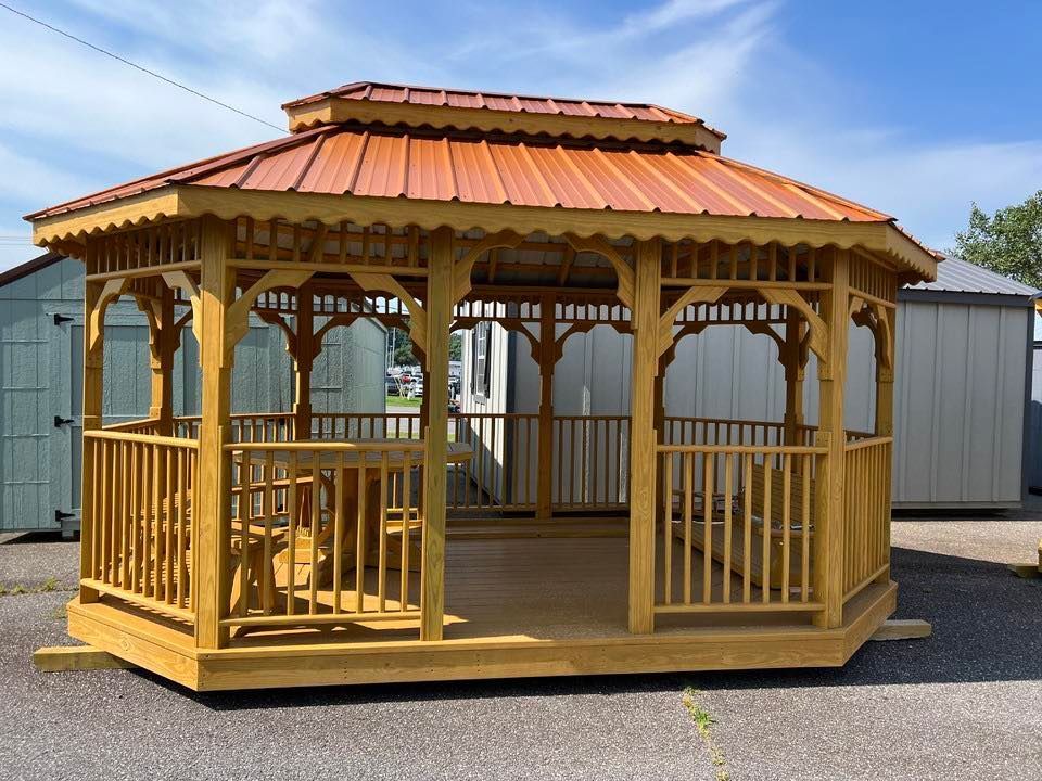 Wooden gazebo with orange metal roof, sitting on a paved lot. A sunny day with clear blue sky.