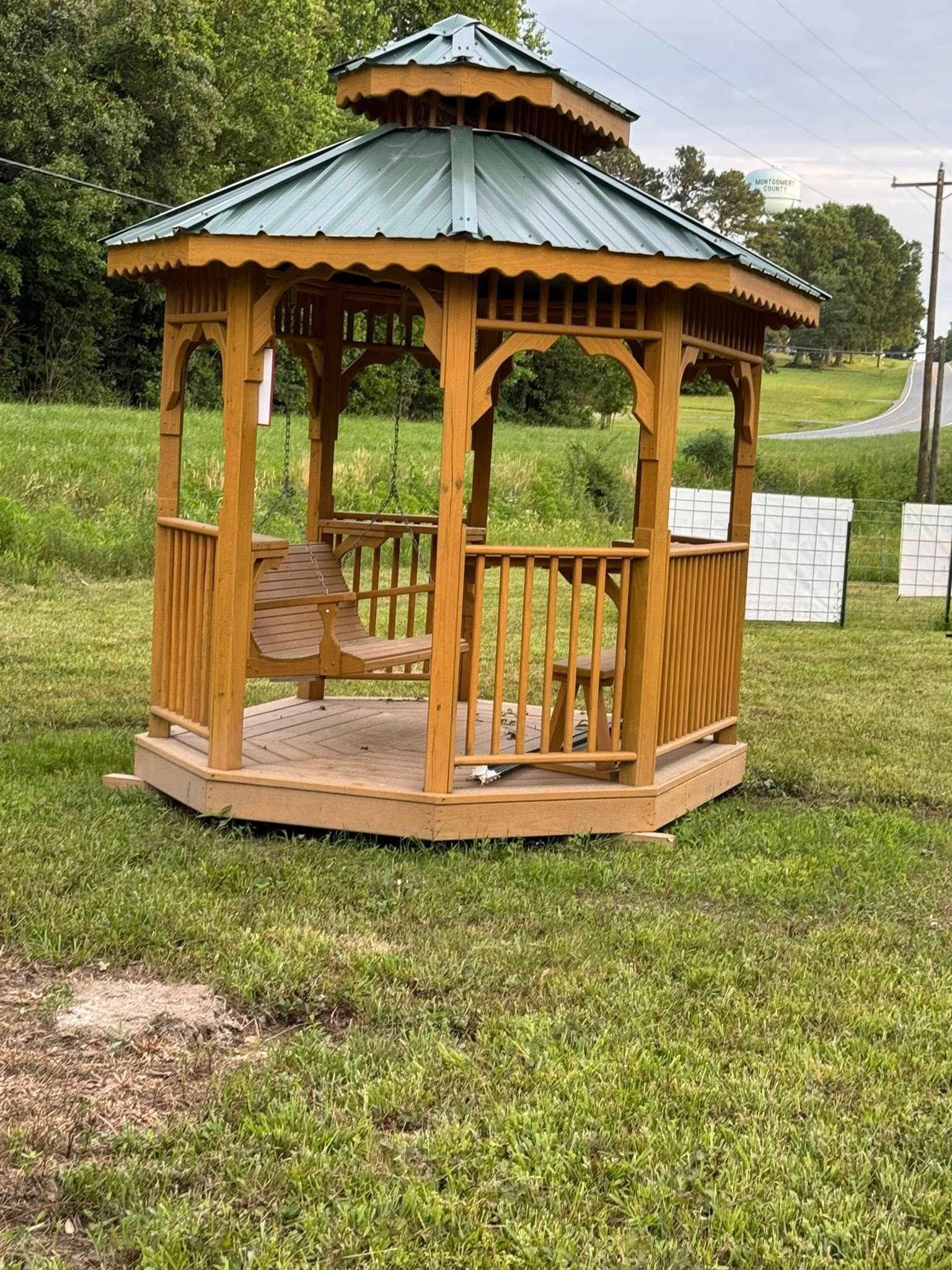 Wooden gazebo with a green metal roof sits in a grassy yard; trees and a road are in the background.